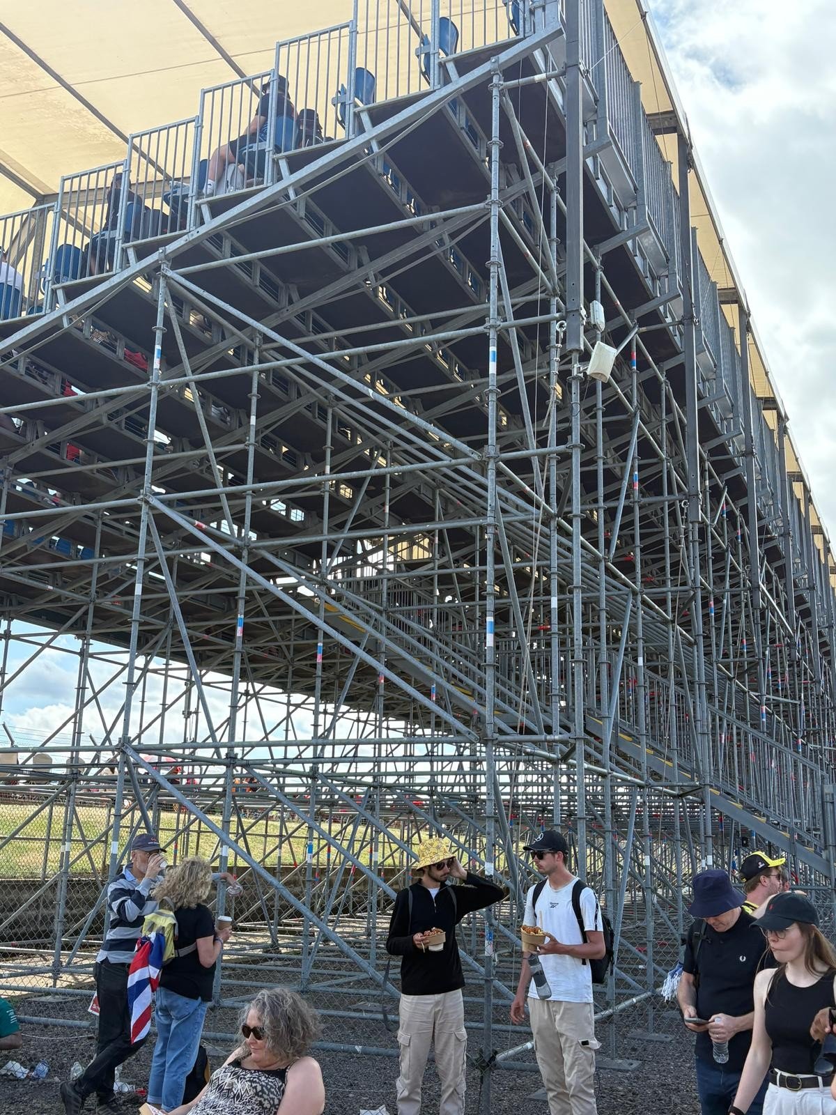 People standing and talking near a large metal scaffolding structure at an outdoor event, with some sitting on the structure.