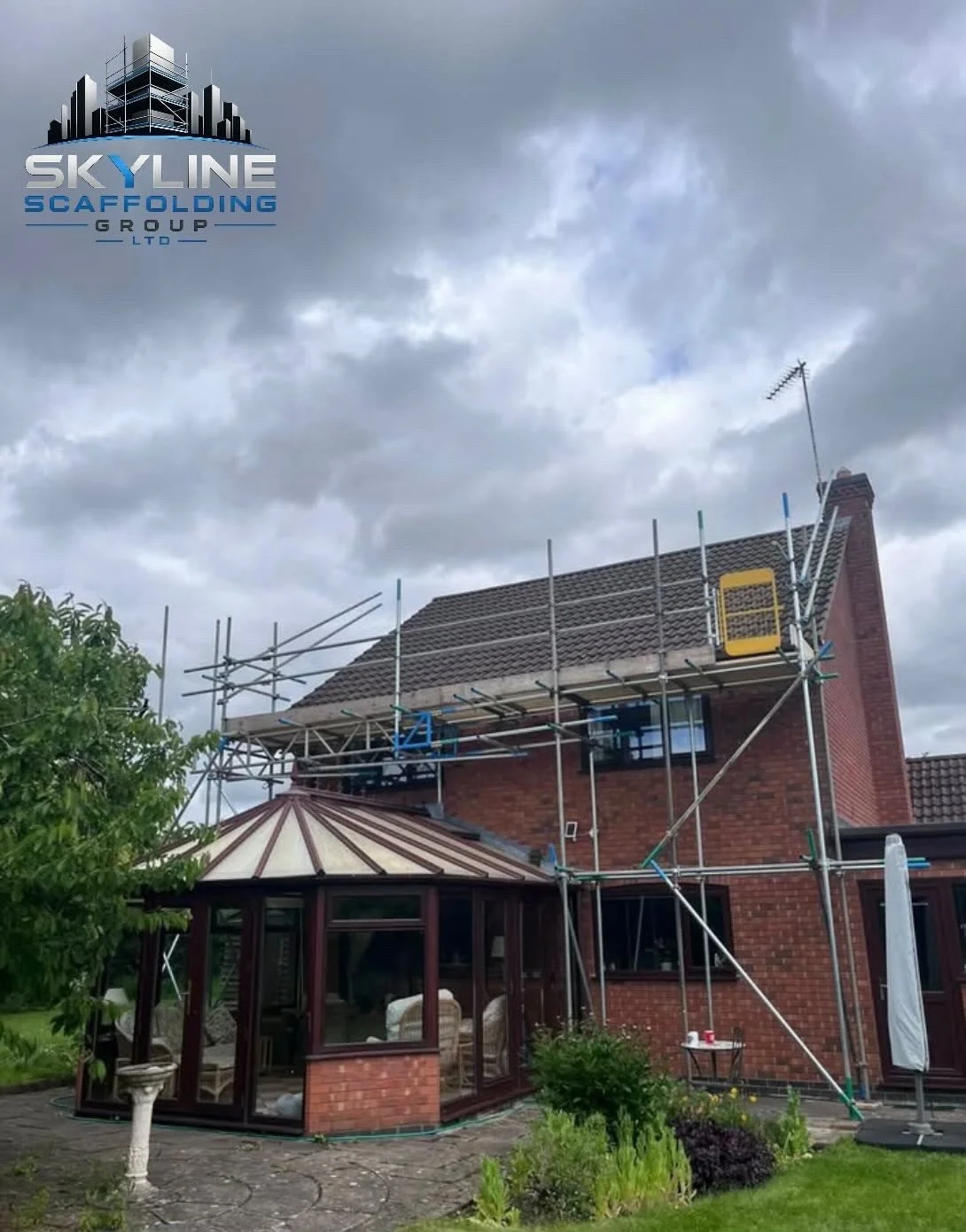 A two-story red brick house with scaffolding on the side, and a conservatory attached to the back. The sky is mostly cloudy.