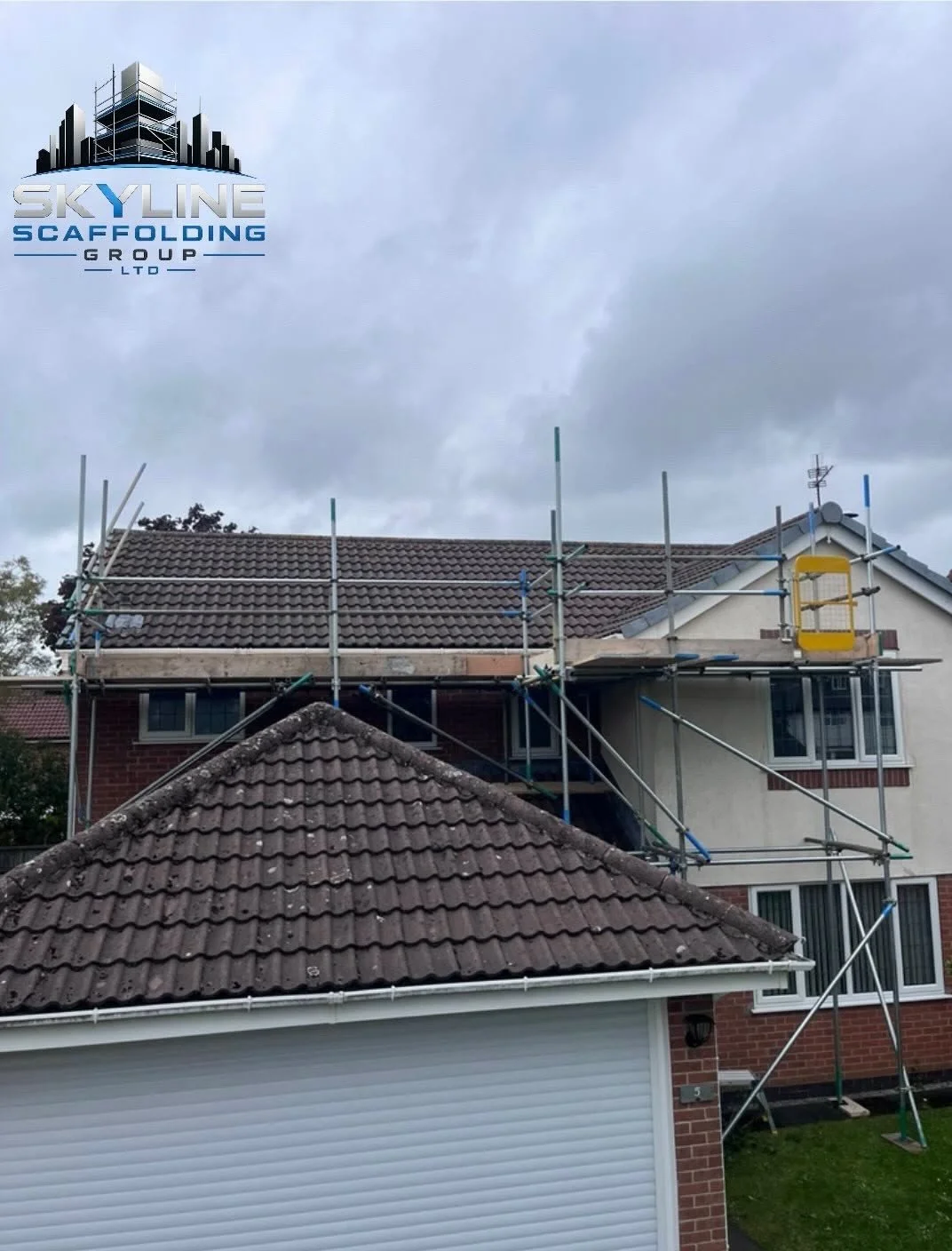 Scaffolding set up around a house with a brown tiled roof, preparing for roof work, under a cloudy sky.