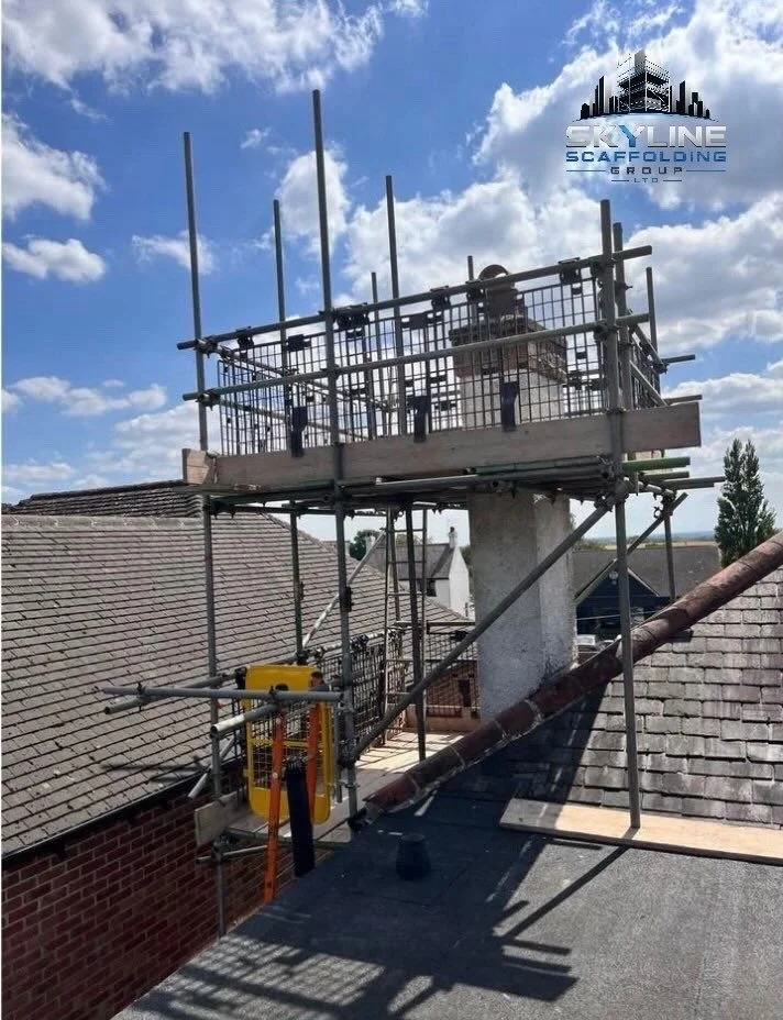 A construction scaffold attached to a residential rooftop with safety railing, some equipment, and an adjacent chimney. Cloudy sky in the background with a logo of Skylines Scaffolding Group in the upper right corner.