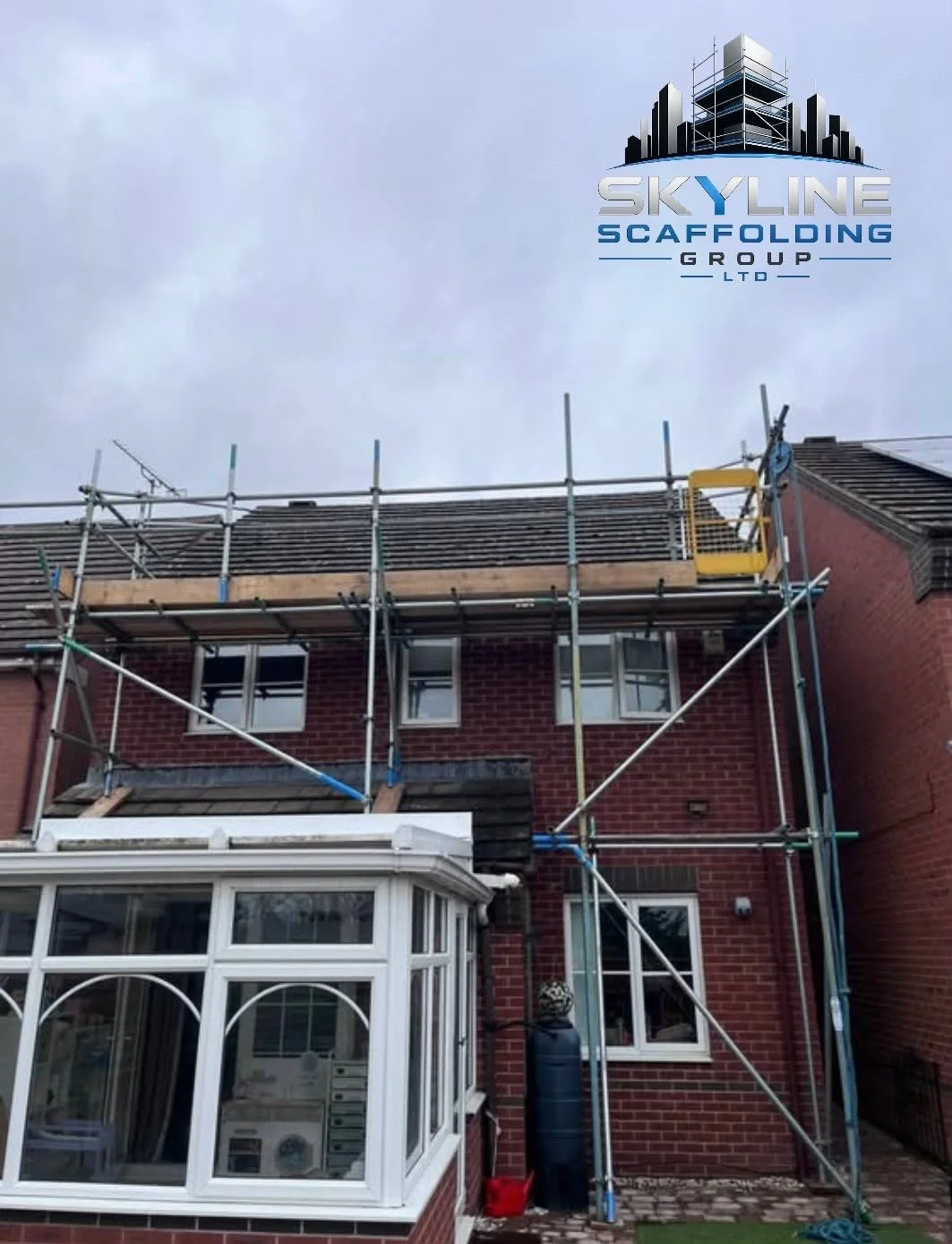 House with scaffolding on the roof, under repair or renovation, with a conservatory in the foreground and cloudy sky above.