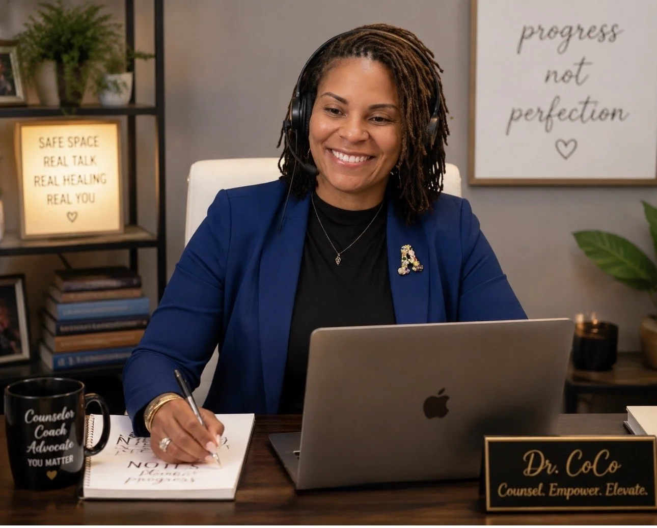 Smiling woman wearing a headset and blue blazer writes beside a laptop at a counseling office desk.