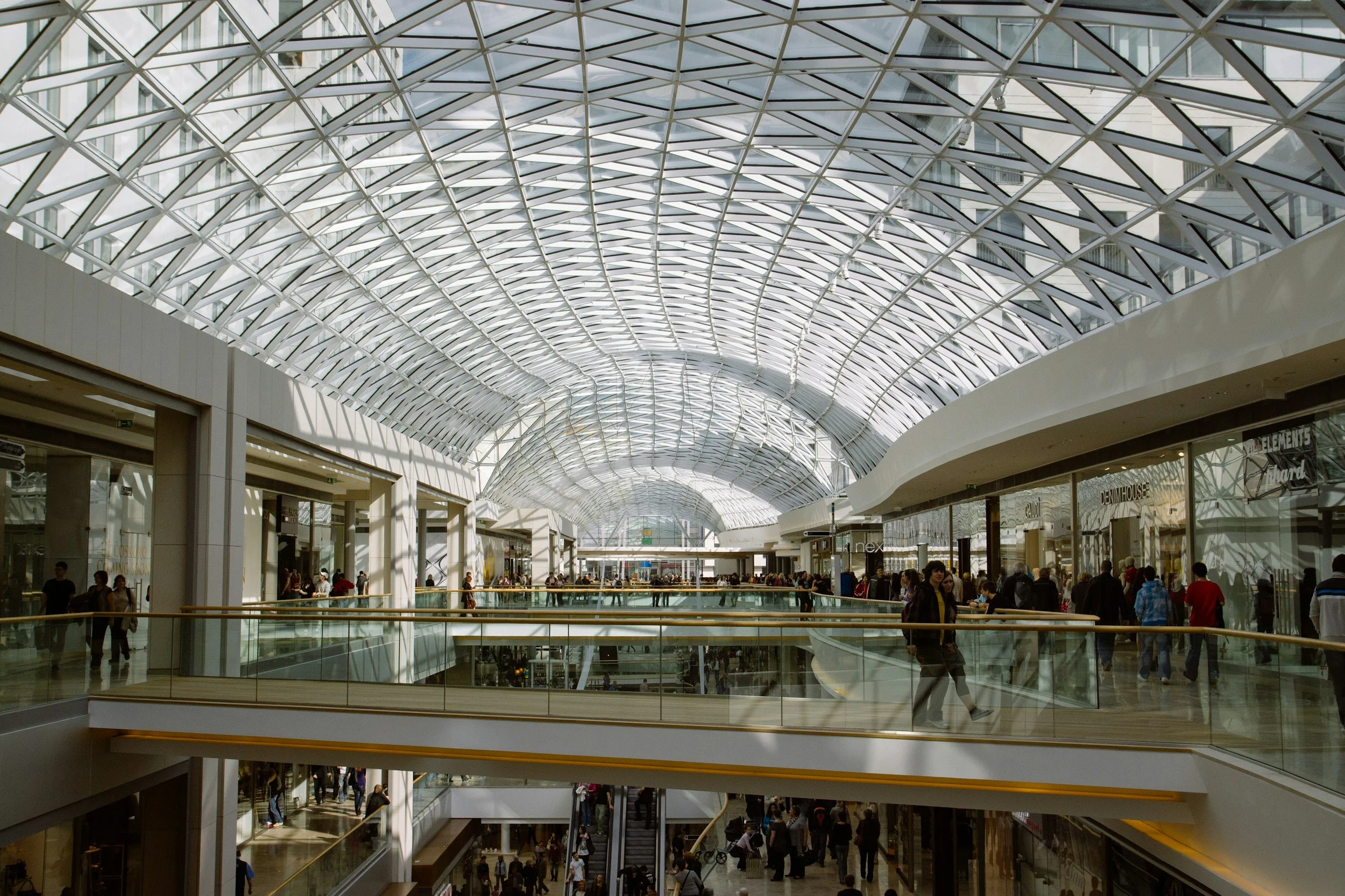Interior of a modern shopping mall with a glass, arched ceiling, multiple levels, and people walking around.