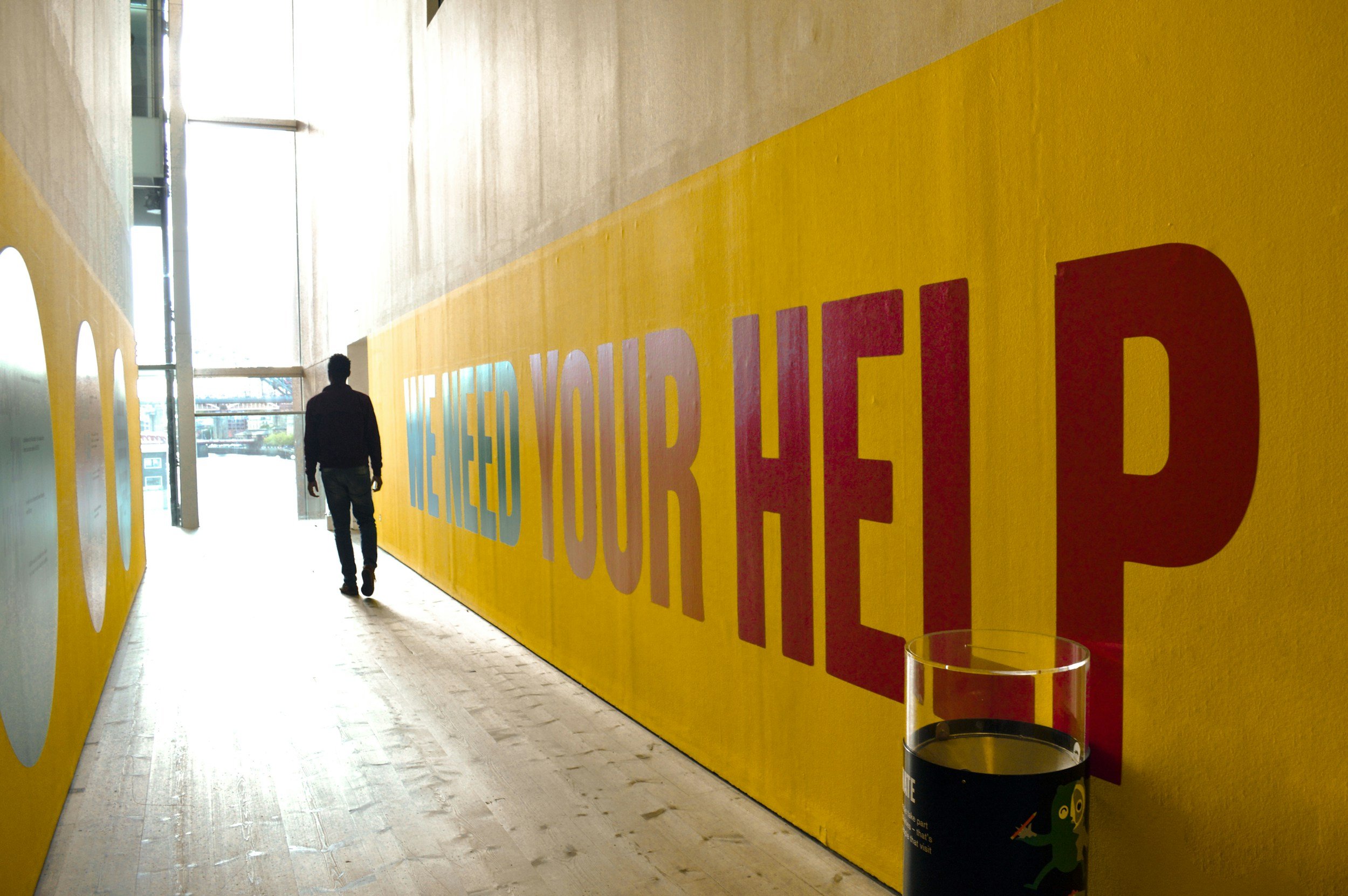 Ein dunkler Mann läuft durch einen Flur mit einer großen gelben Wand, auf der teilweise bunte Buchstaben zu sehen sind. Am Ende des Flurs sind Fenster, durch die Tageslicht hineinströmt.