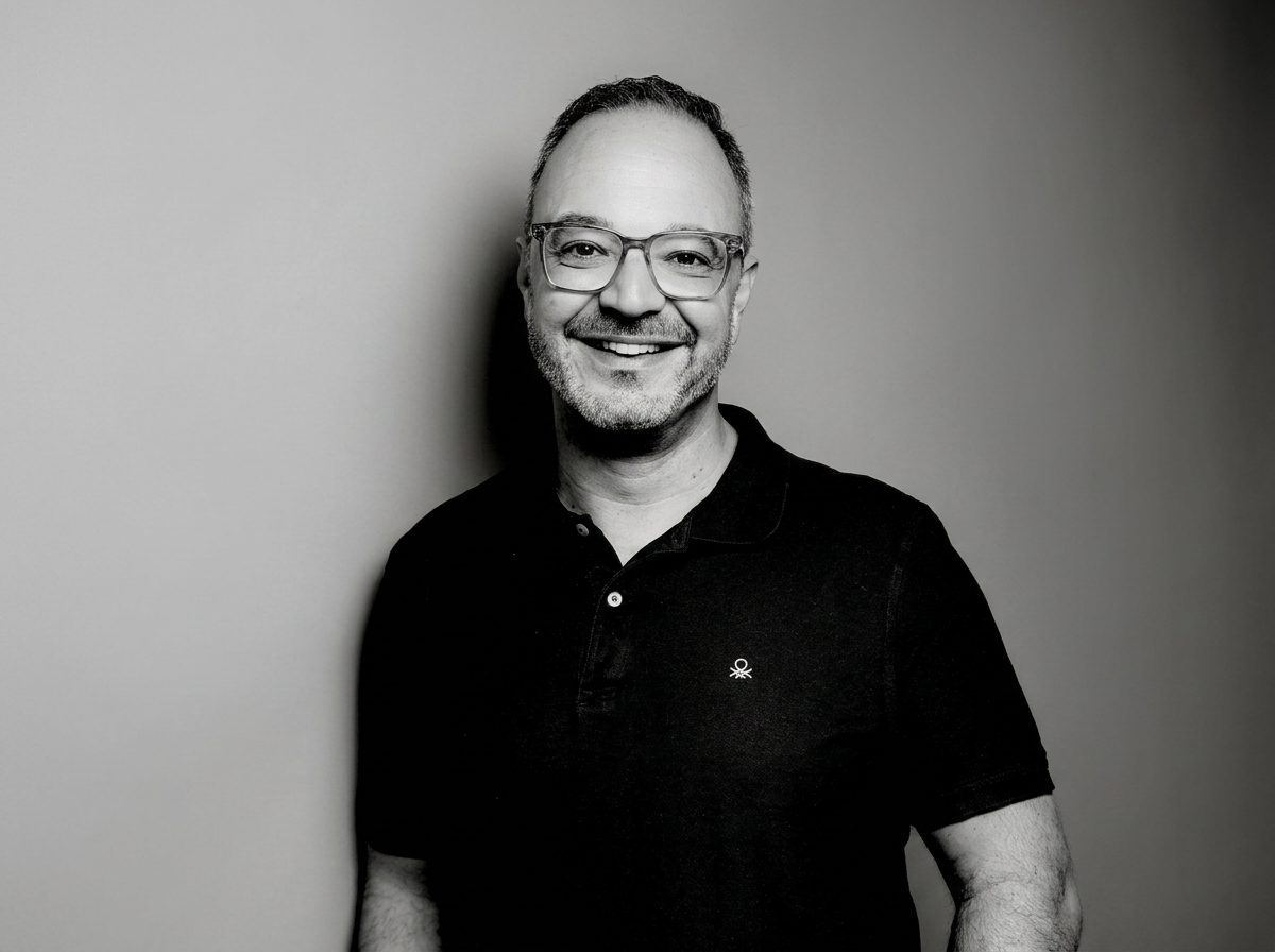 Lächelnder Mann mit Brille, grauem Bart, schwarz Polo-Shirt, gegen hellgrauen Hintergrund stehend.