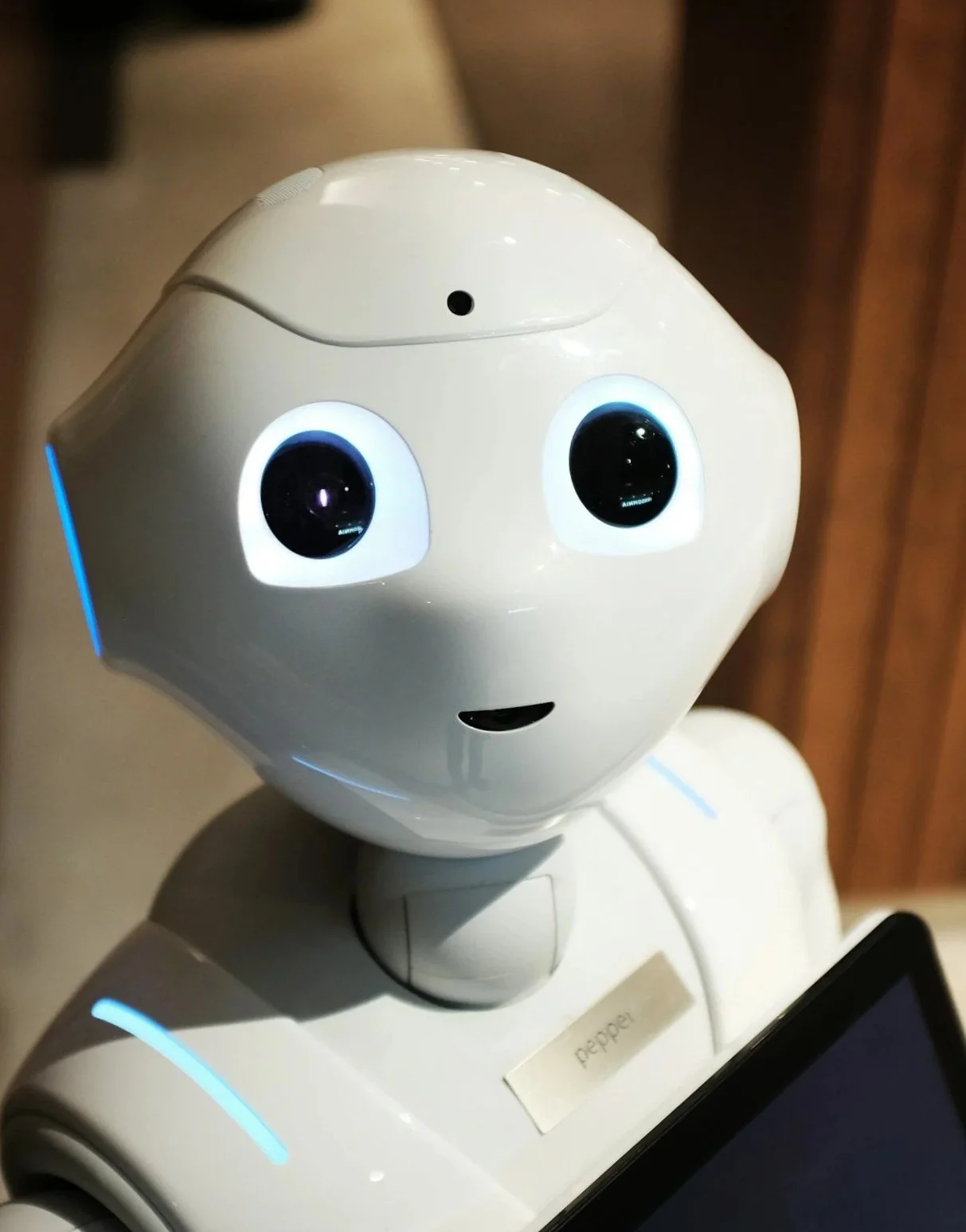 Close-up of a white humanoid robot with large black eyes, a small mouth, and a smooth rounded head, against a wooden background. Explore Kamtron's solutions and services
