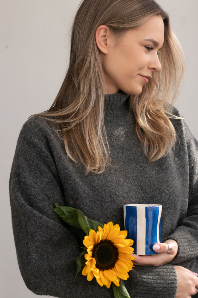 A woman with long blonde hair wearing a gray sweater holding a sunflower and a blue-and-white striped mug, smiling while looking to her right.