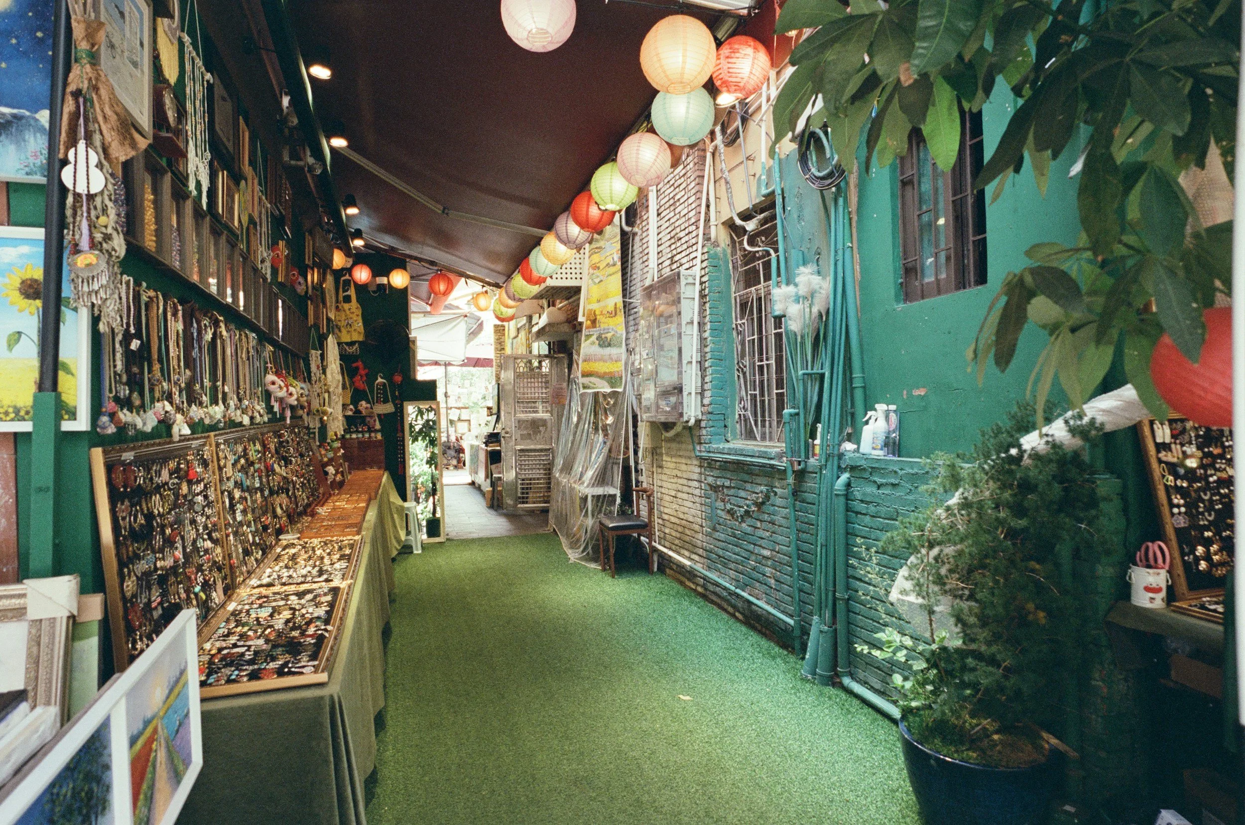 An alleyway with decorative paper lanterns hanging above and a green artificial grass floor. The left side contains a stall displaying various jewelry and accessories, while the right side has a large potted plant and a green painted wall. The alley is partially covered with a brown ceiling and appears to be inside or part of a marketplace or shopping area.