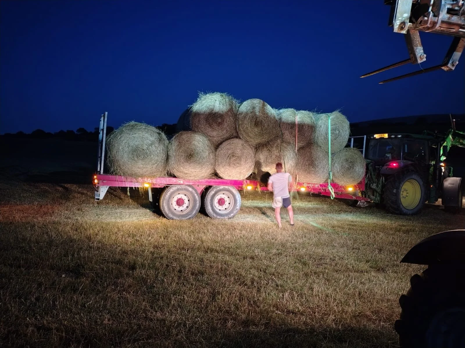 A man is standing next to a flatbed trailer loaded with hay bales. The trailer is attached to a green tractor, and the scene is illuminated at night with a dark blue sky in the background.