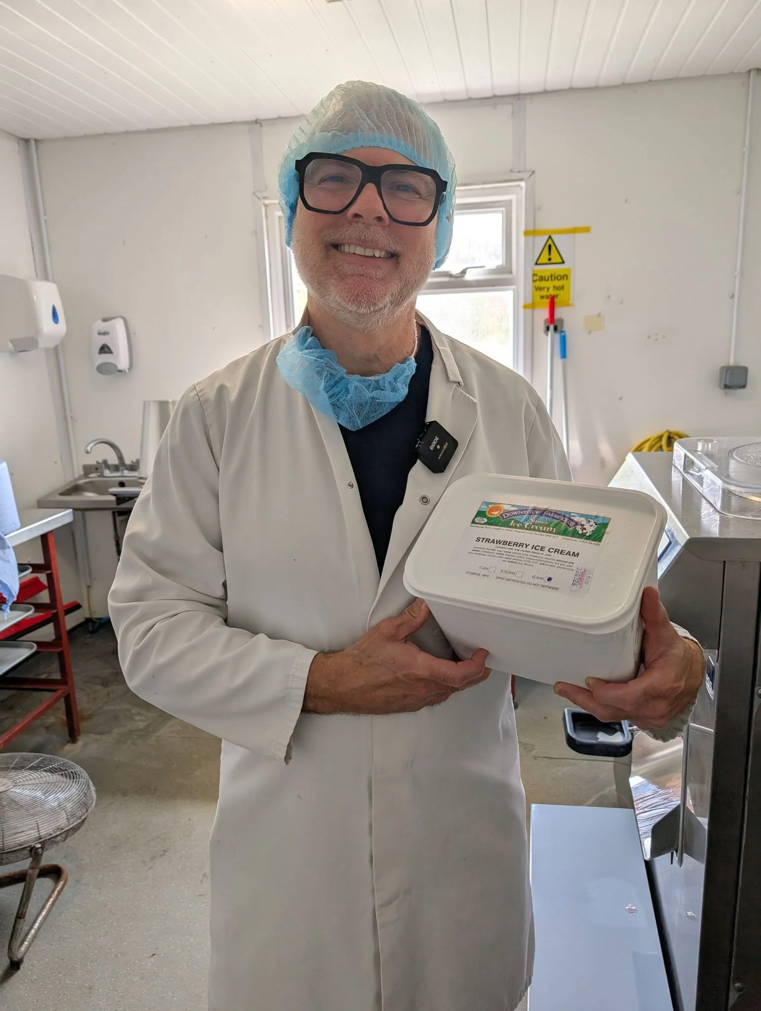 A man with glasses and a beard smiling while holding a container of strawberry ice cream in a commercial kitchen.