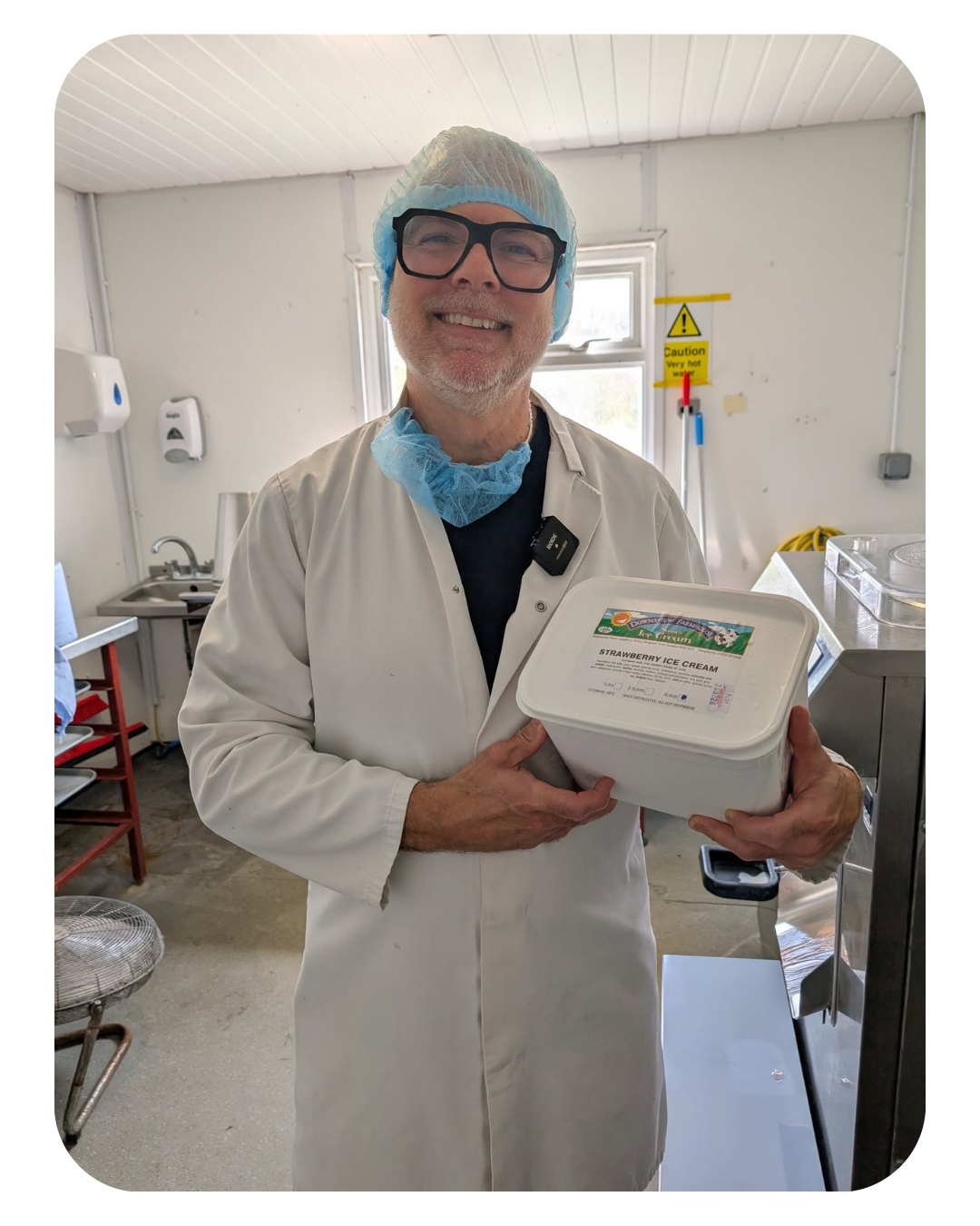 A man in a white lab coat, wearing glasses, a hairnet, and a blue face mask pulled down, is smiling and holding a container of strawberry ice cream in a commercial kitchen or ice cream factory.
