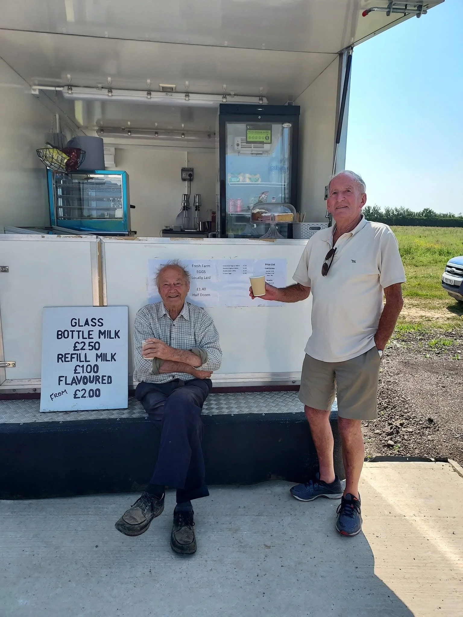 Two elderly men in casual clothing standing and sitting in front of a mobile milk stand with signs displaying prices for glass bottle milk, refill milk, and flavored milk, set outdoors on a sunny day.