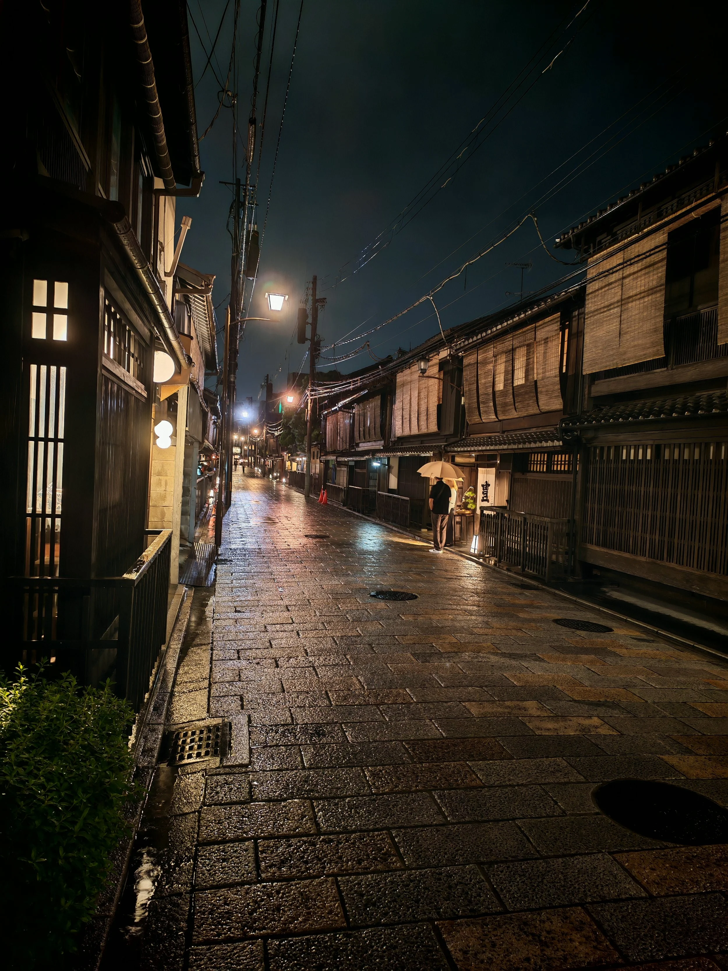 A rainy night scene of a narrow street in Japan with traditional wooden buildings, illuminated by street lamps, and a person holding an umbrella walking.
