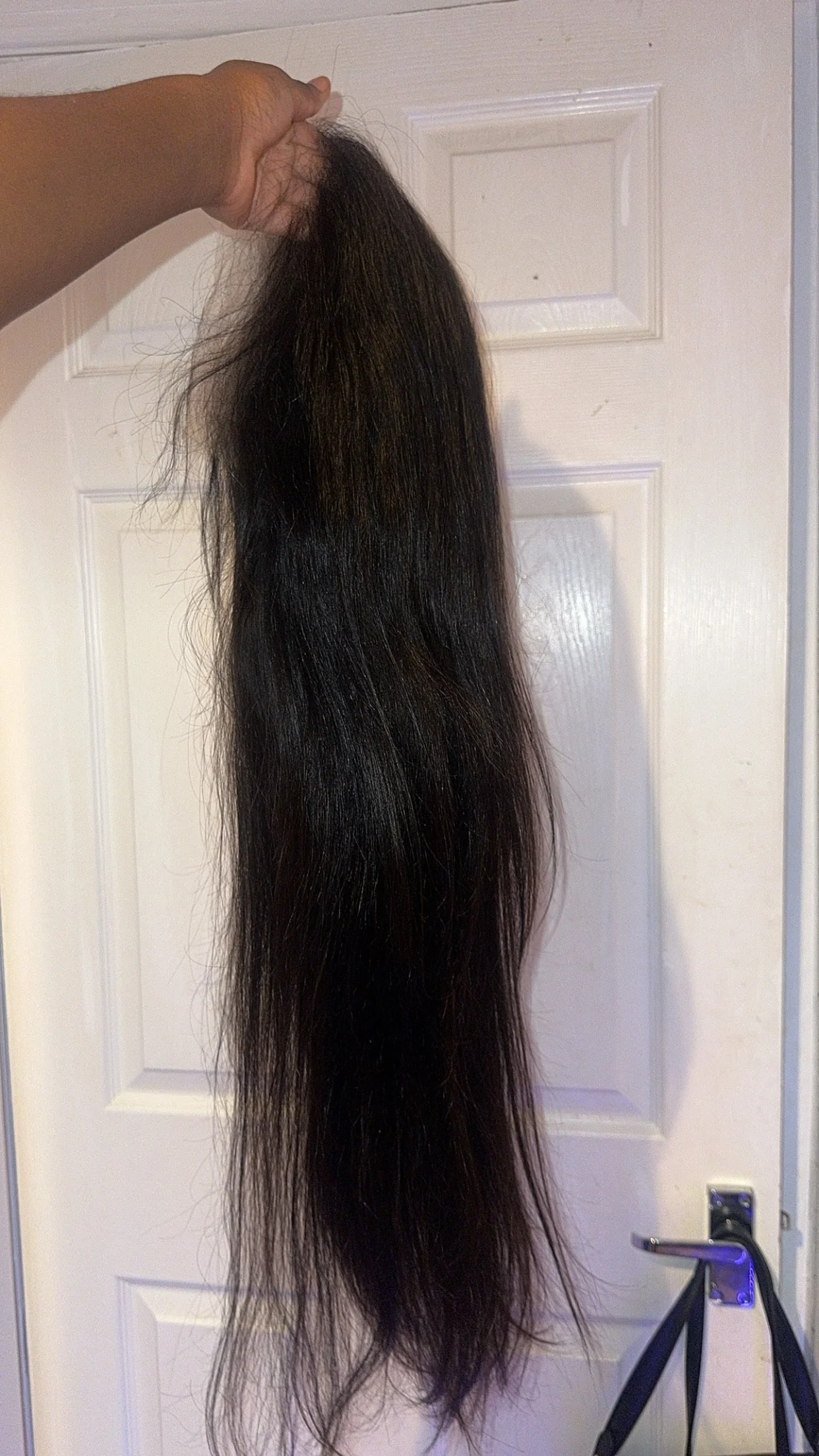 A long, thick, black hair extension or wig hanging from a hand against a white door with a silver door handle.