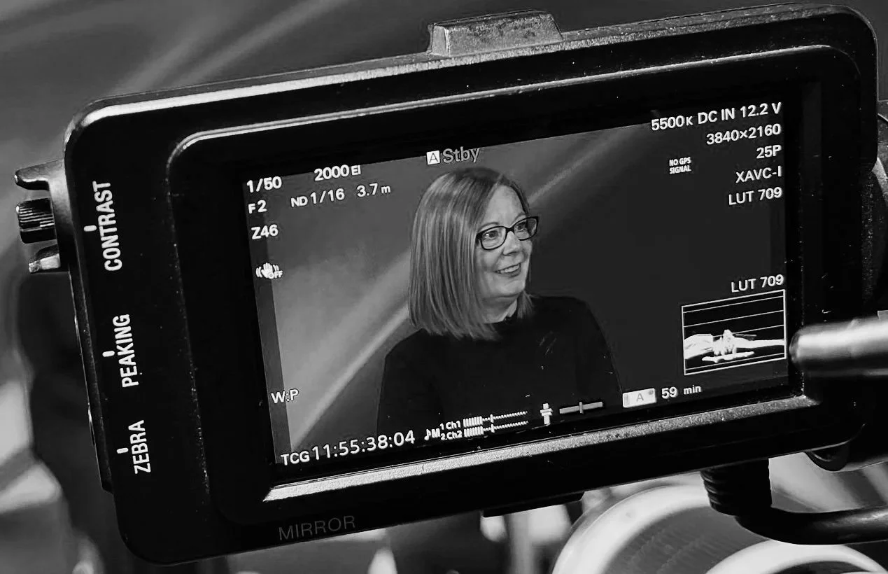 A television camera captures an image of a woman with shoulder-length hair and glasses, smiling and wearing a dark top, during a recording or broadcast.