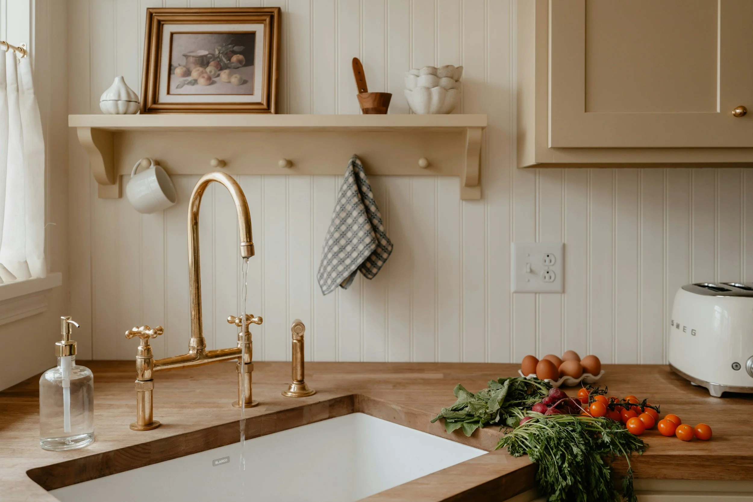 A kitchen sink with a copper faucet, a bottle of hand soap, a bunch of fresh vegetables including tomatoes and leafy greens on the wooden countertop. A wall shelf with art, a cutting board, a white bowl, and a ceramic pumpkin. Part of a window with white curtains, a beige kitchen cabinet, and a white blender.