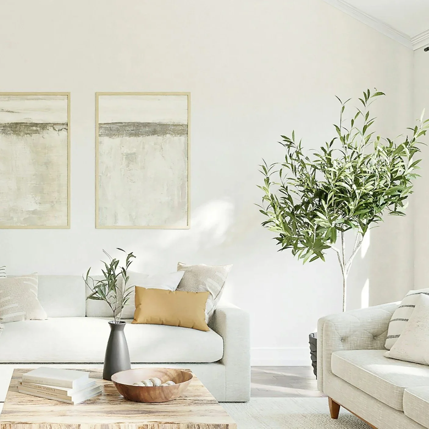 A bright, minimalist living room with white walls, a white sofa with beige and white pillows, a large potted plant, two framed abstract paintings, and a rustic wooden coffee table with books, a bowl, and a small vase with greenery.