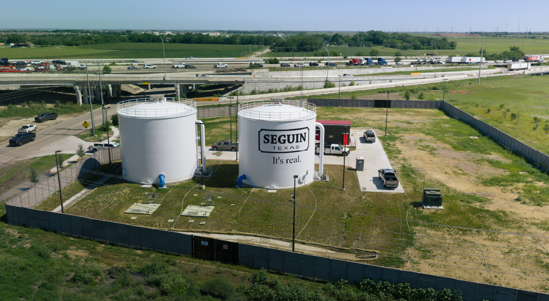 Two large white storage tanks with the sign 'Seguin Texas, It's real.' on one tank, surrounded by a fence, a parking lot with several vehicles, and a power line. Highway with traffic in the background.