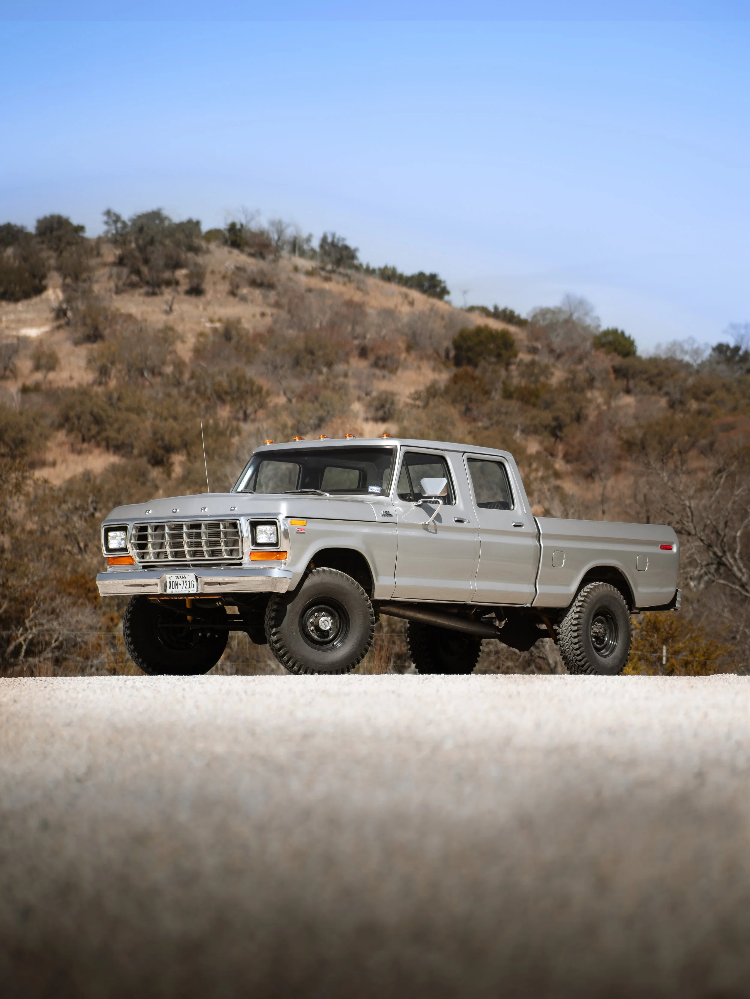 A silver vintage pickup truck parked on a gravel surface with a hillside covered in trees and shrubs in the background under a clear blue sky.