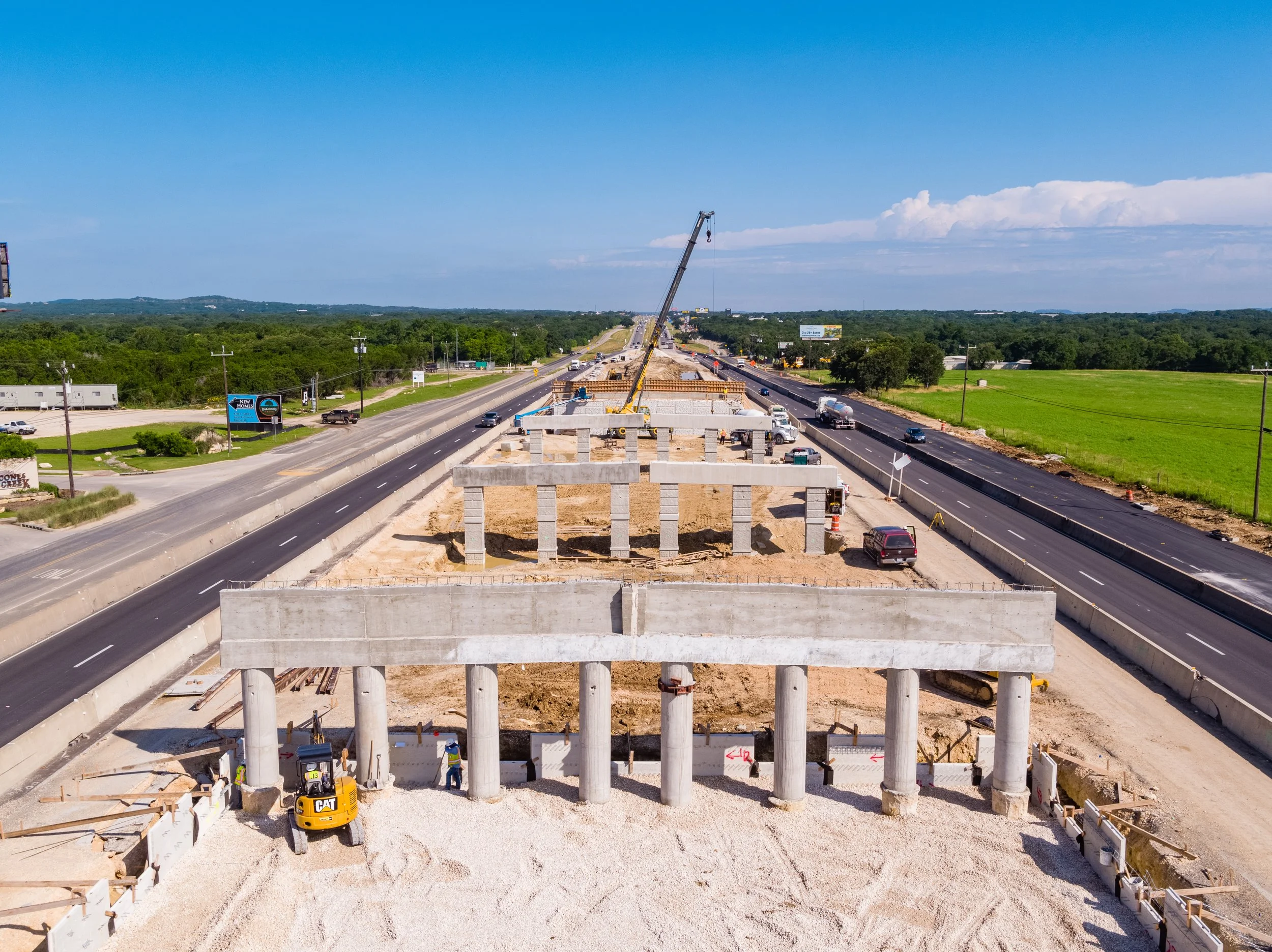 Construction site on a highway with large concrete pillars being built and a crane lifting materials, vehicles on the road, green fields, and a blue sky with clouds.