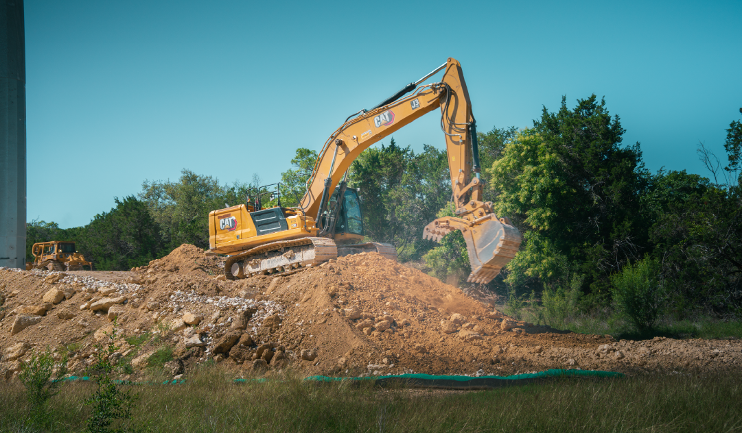An orange excavator on a mound of dirt working on a construction site, with trees in the background under a clear blue sky.