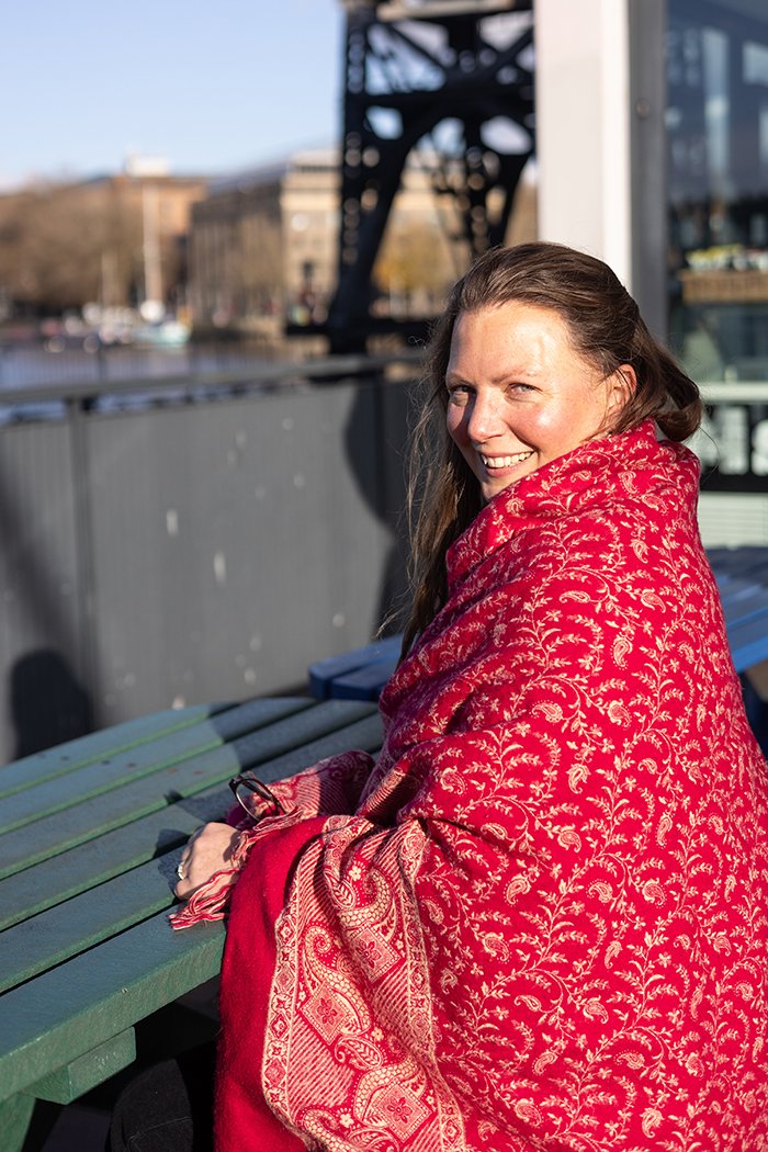 Photo of Lesley wrapped in a red shawl, sitting in the sunshine on Bristol Habourside.
