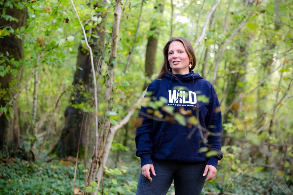 Photo of Lesley, standing in the woods wearing a hoodie with the word 'wild' on it. She is healthy and happy to be outside.