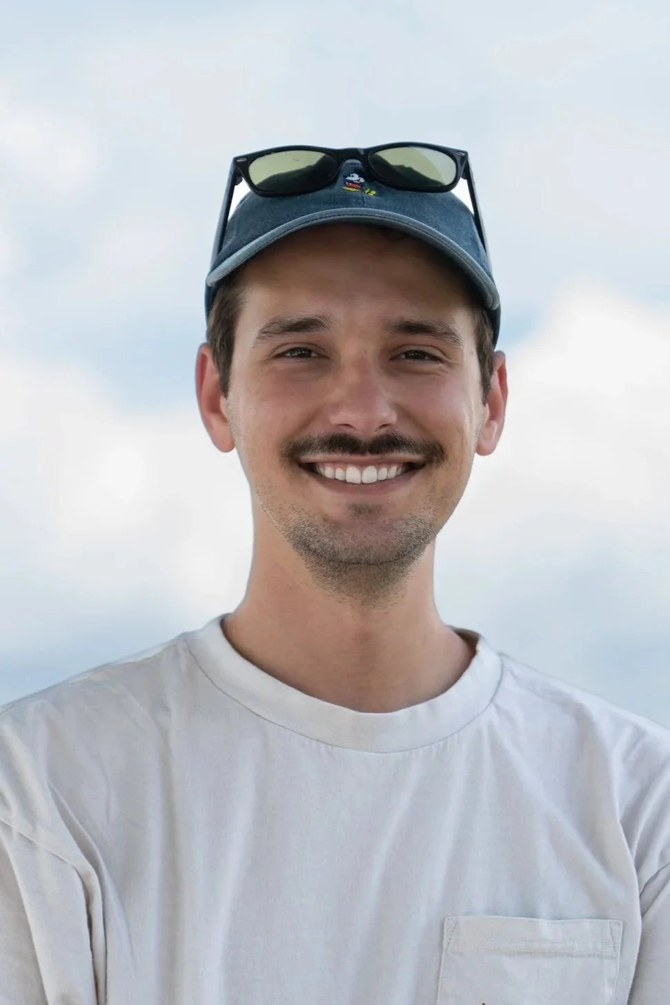 A young man with a mustache and beard smiling, wearing a gray baseball cap with sunglasses on top, a white t-shirt, and standing outdoors with a cloudy sky in the background.