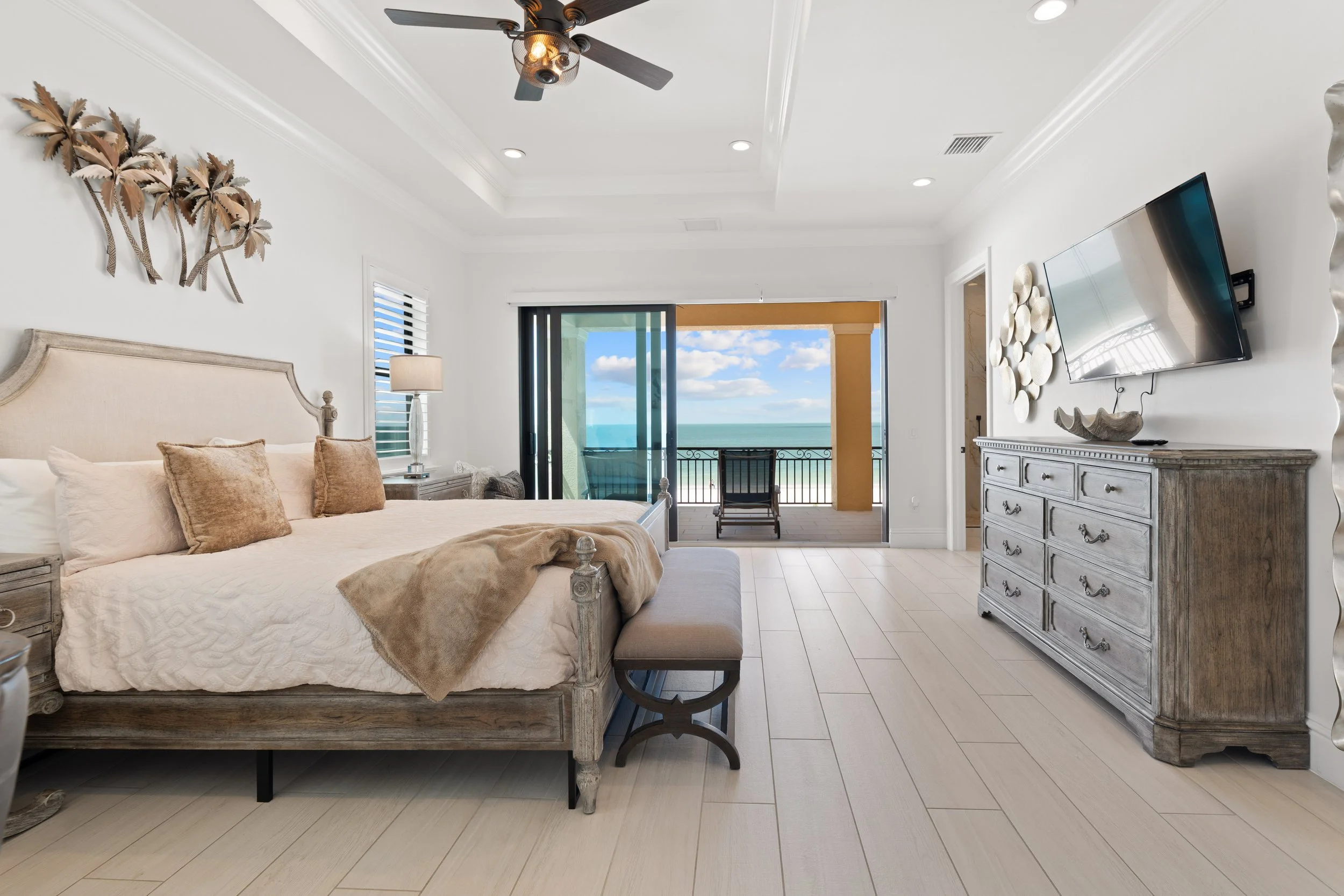 A bedroom with a wooden bed, beige bedding and pillows, a side table with a lamp, a sliding glass door leading to a balcony with ocean view, a wall-mounted TV, a dresser, and ocean scenery visible outside.