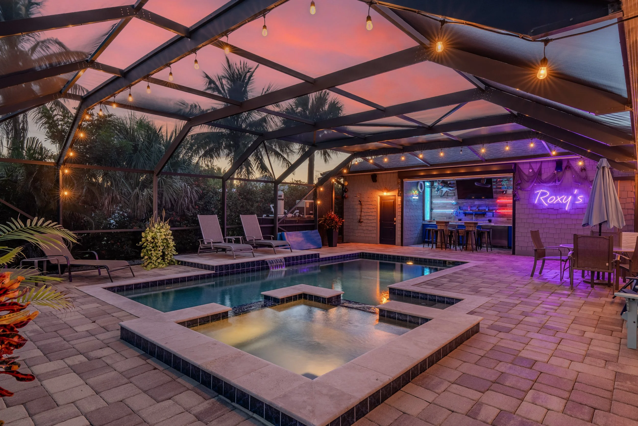 A backyard view during sunset with a screened-in patio area featuring string lights, a pool, lounge chairs, a bar area, and palm trees in the background.