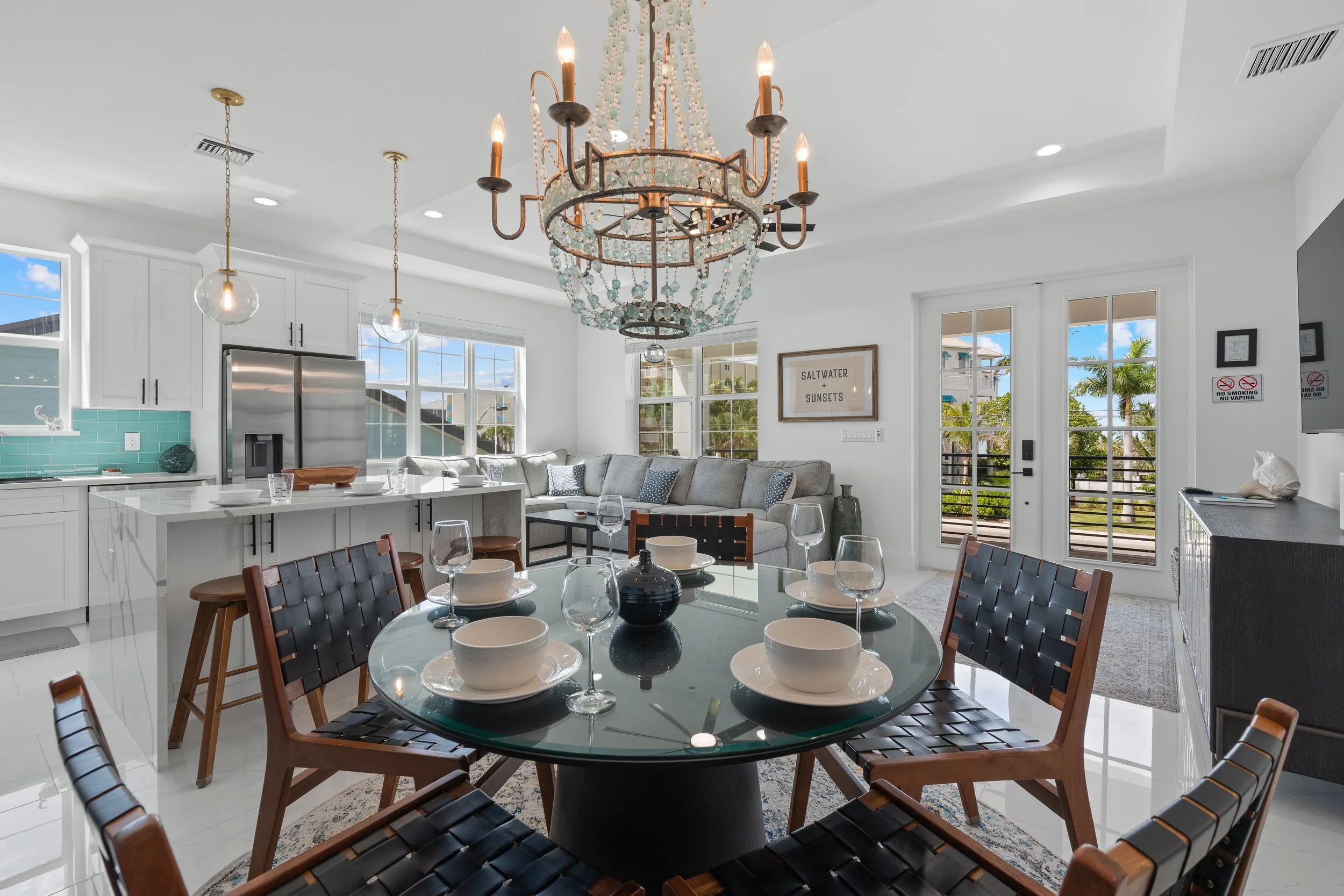 Bright, modern open-concept kitchen and dining area with a large chandelier, a round glass dining table set with white dishes and glasses, dark wood chairs, white cabinetry, stainless steel refrigerator, light blue backsplash, large windows, and a living space with a gray sectional sofa and framed wall art.