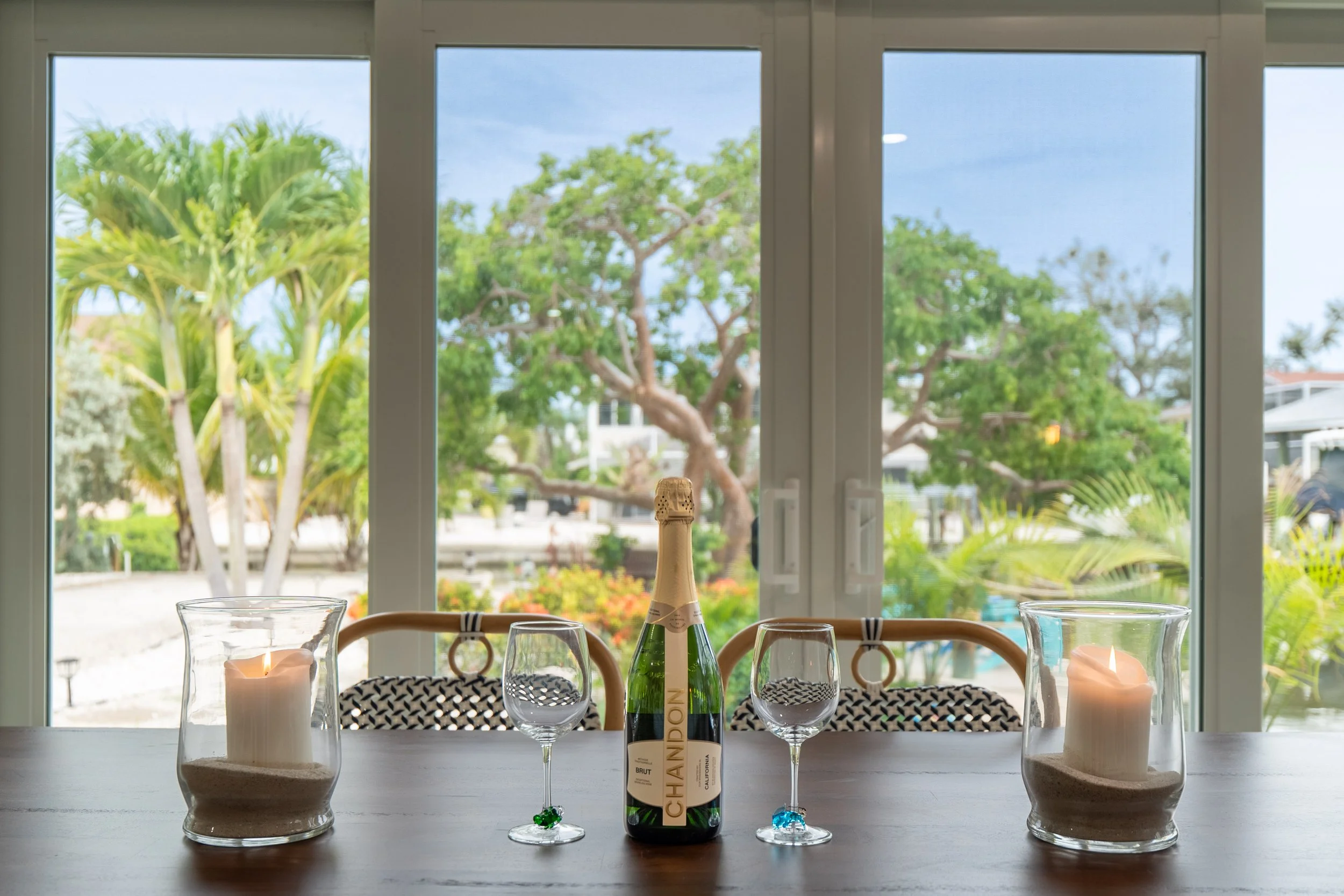 Table set with candles, glasses, and a bottle of Champagne in front of large windows showing trees and a backyard.
