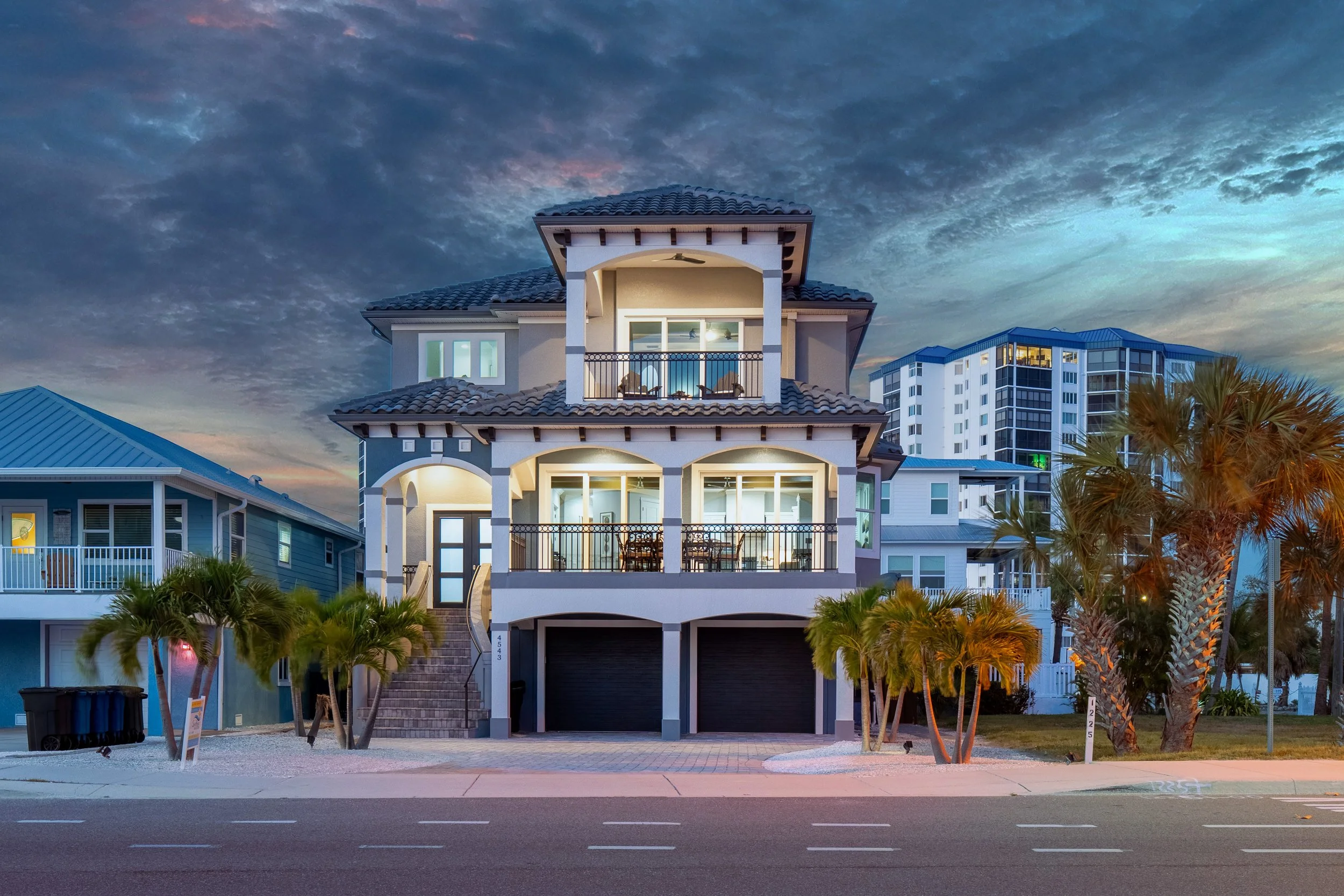 A modern three-story house with a tiled roof and expansive balconies, situated in a neighborhood with other tall buildings, palm trees, and a partly cloudy sky at dusk.