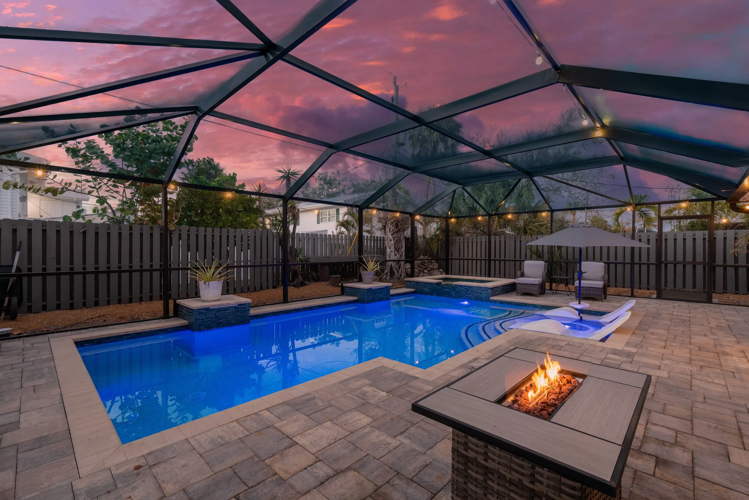 A backyard swimming pool area during sunset with a screened enclosure, outdoor furniture, string lights, and a small fire pit.