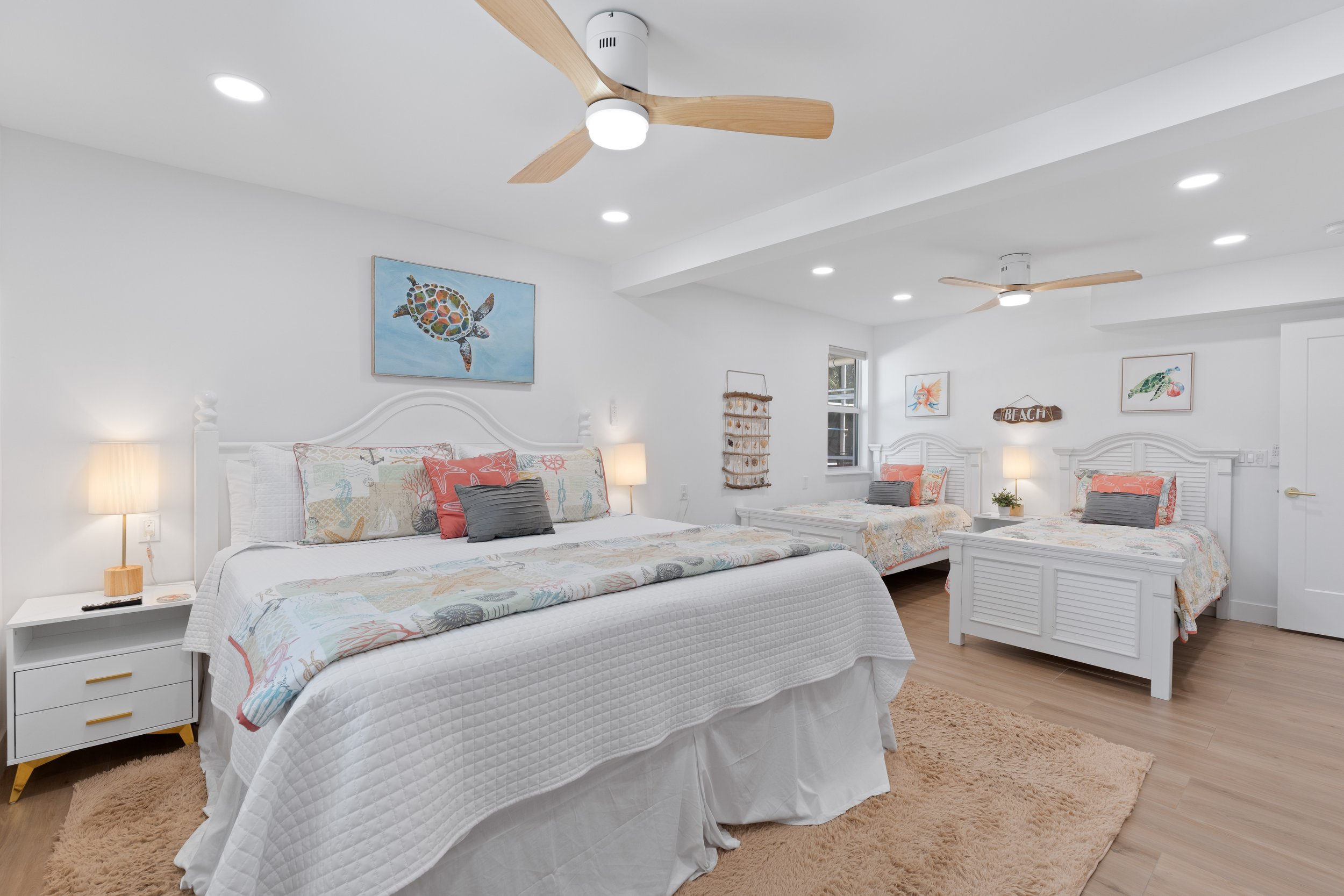 A bright bedroom with two beds covered in white bedspreads with colorful ocean-themed pillows, wooden side tables with lamps, white walls with sea turtle and beach-themed artwork, ceiling fans, and light-colored wooden flooring with a beige rug.