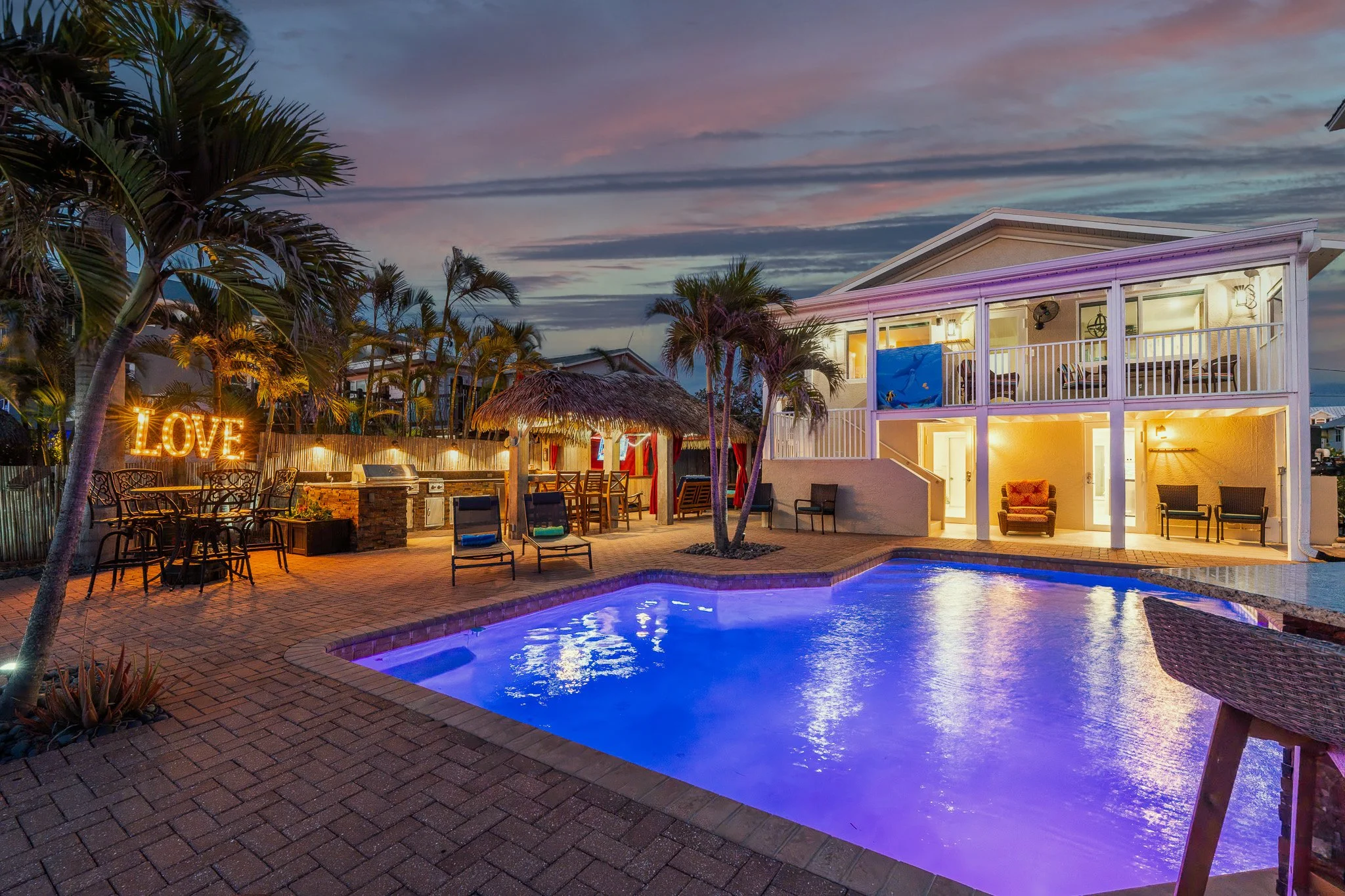 A backyard with a swimming pool illuminated at night, surrounded by lounge chairs, a tiki hut, and palm trees. There is a two-story house with a balcony and lit interior. The sky is dark with colorful clouds, and decorative lights spell out 'LOVE'.