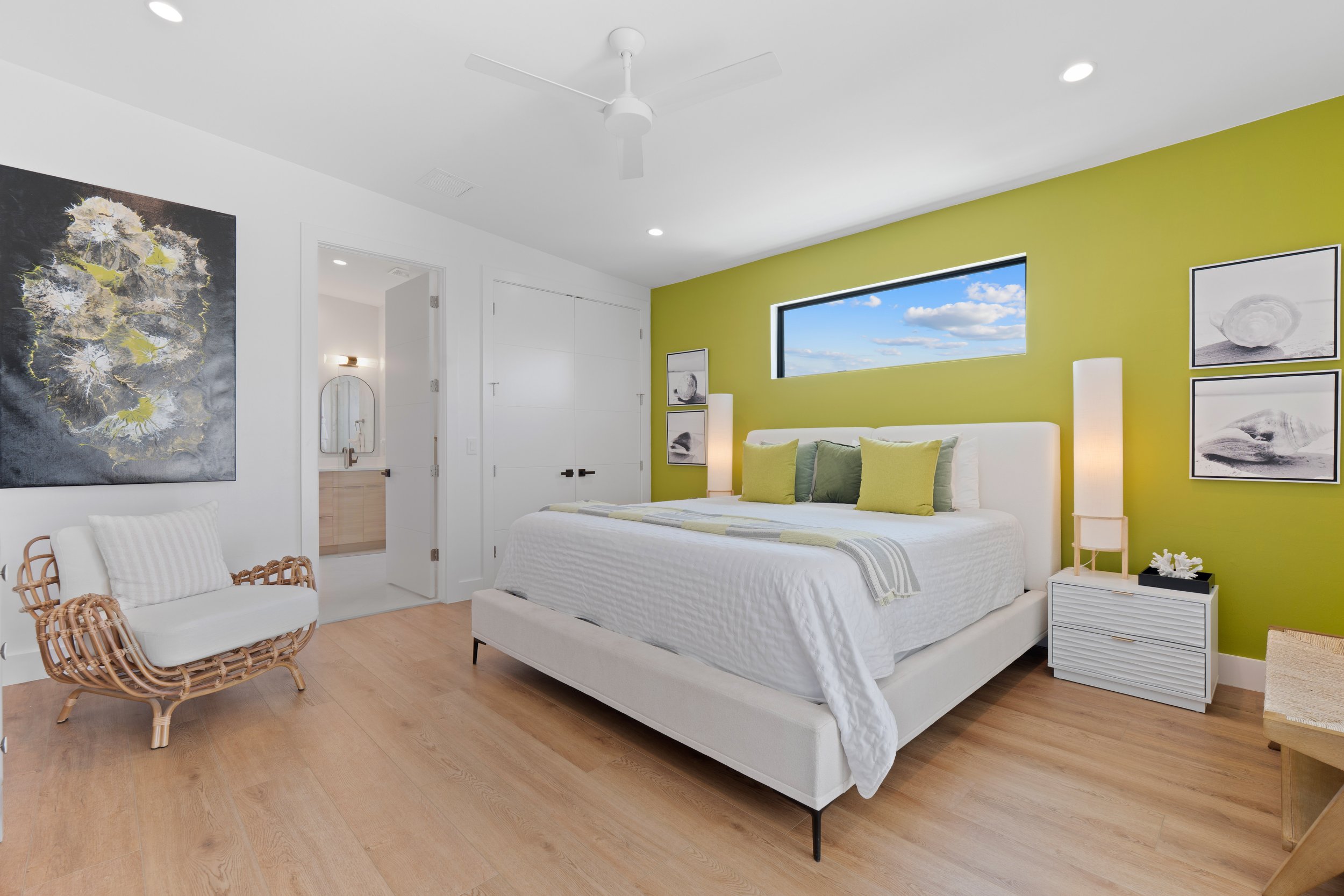 A modern bedroom with a green accent wall, white bed with green pillows, artwork on the walls, and a window showing a blue sky with clouds.