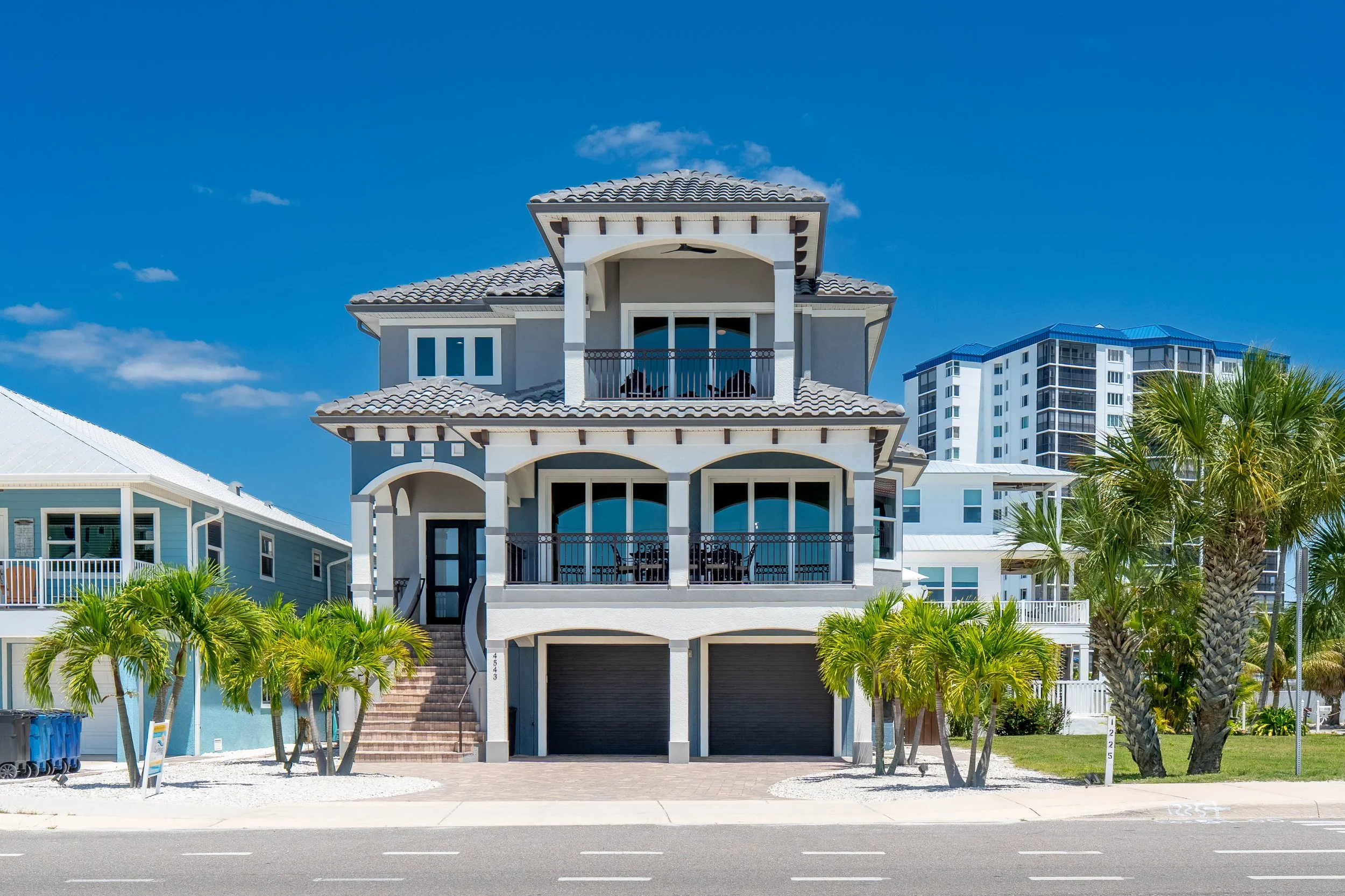 A beach house with three levels, balconies, and a garage beneath, surrounded by palm trees, with a few high-rise buildings in the background, under a blue sky with some clouds.