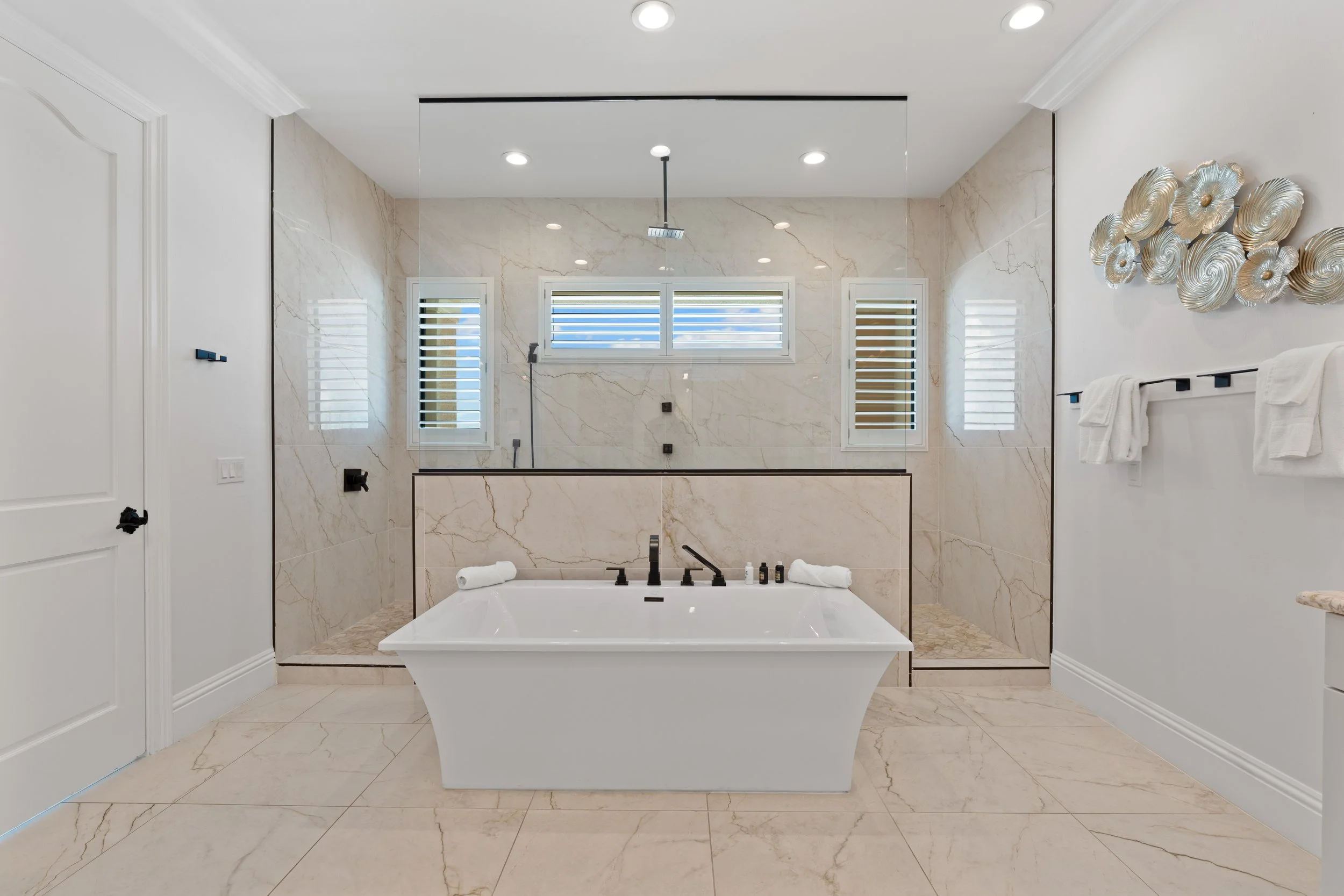 Modern bathroom with a bathtub, marble walls, and three windows with shutters. Decorative wall art is on the right, and towels are on a rack. Recessed ceiling lights illuminate the space.
