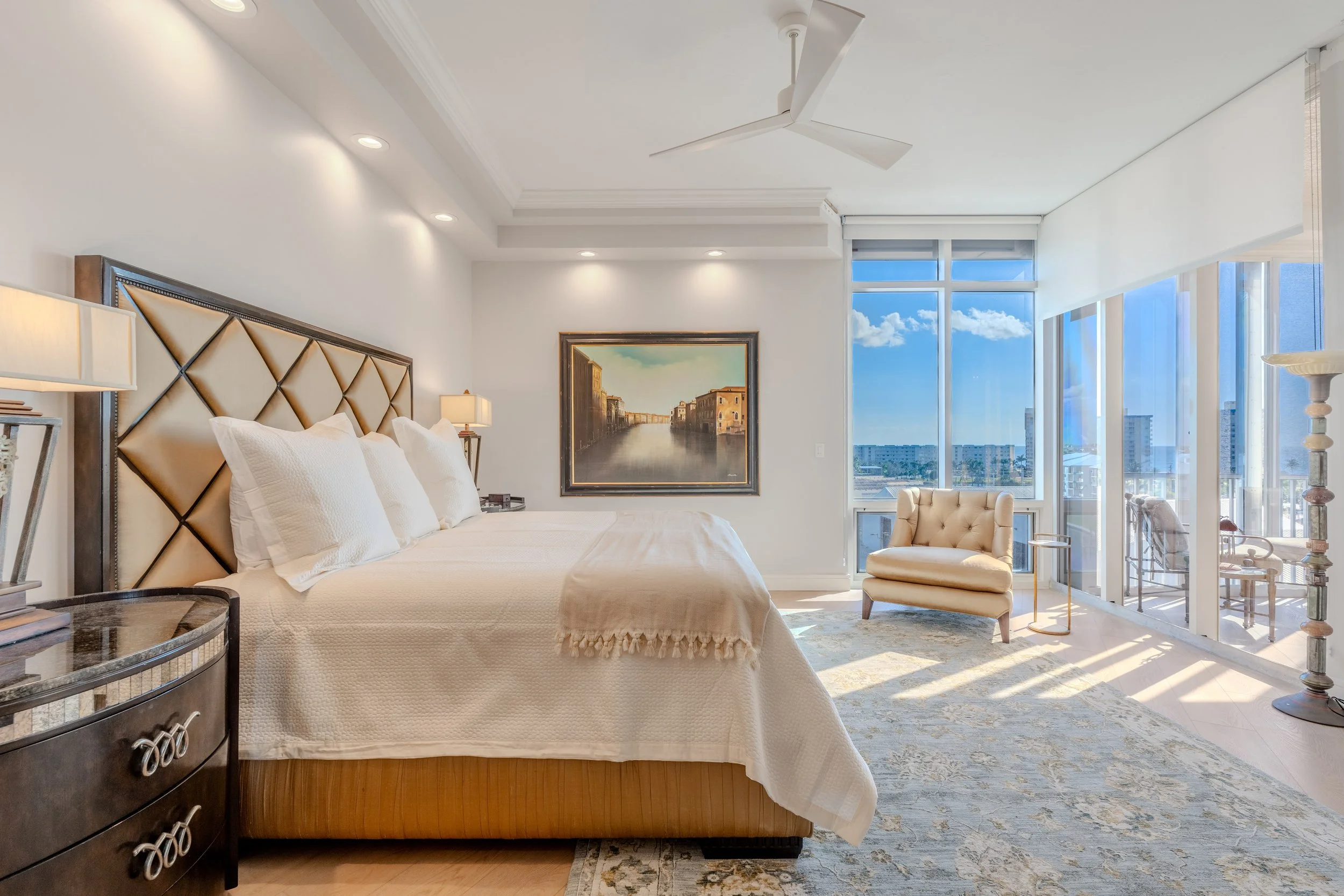 A bright bedroom with large windows showing a city view, a palm tree, and blue sky with clouds. The room has a king-sized bed with a beige headboard and white bedding, with matching lamps and artwork on the walls. There is a cream armchair in the corner and a blue and beige patterned rug on the floor.