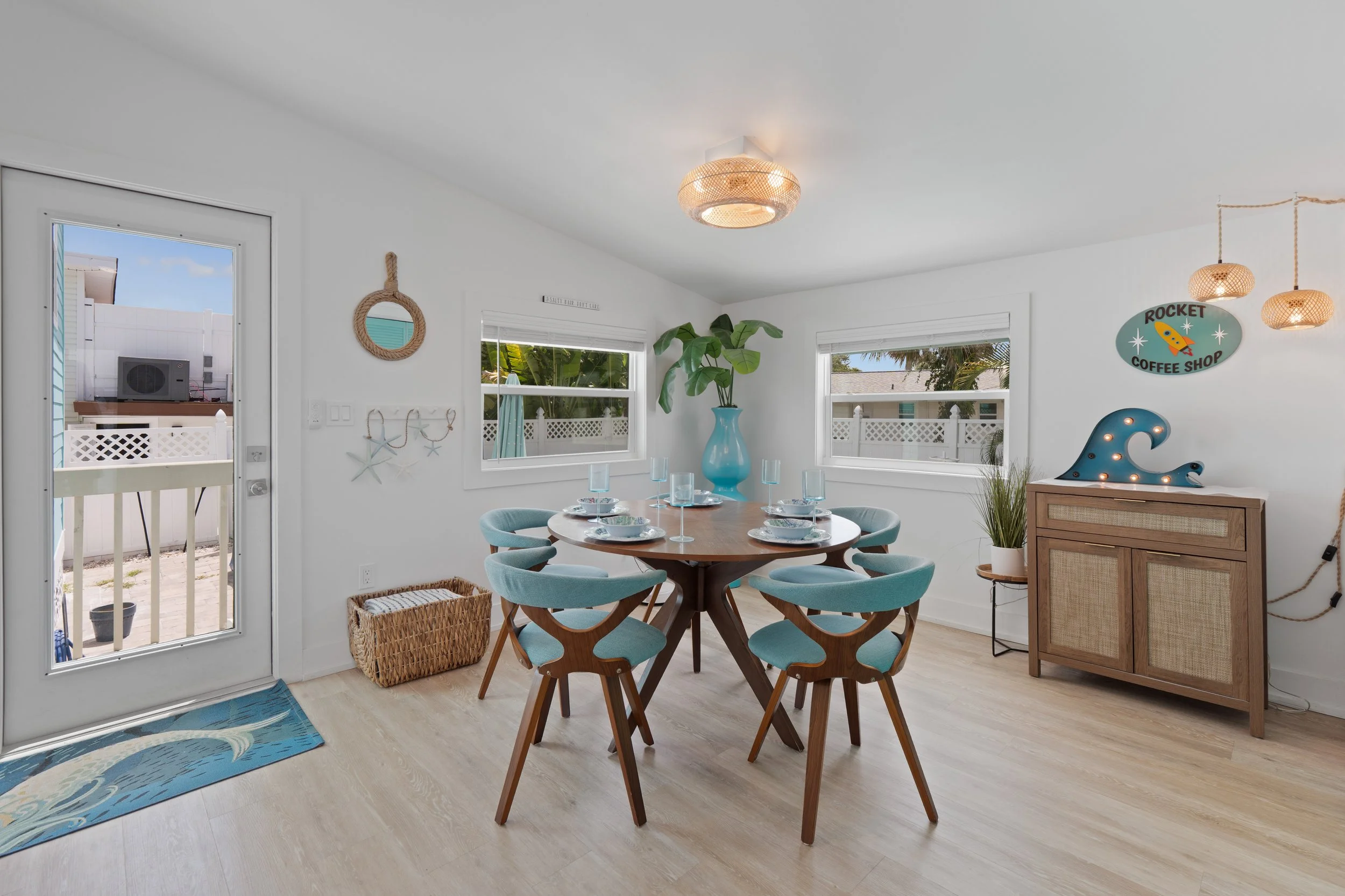 Dining area with round wooden table set with plates, glasses, and utensils, surrounded by leaving room with light-colored flooring, lots of natural light from windows and a door leading outside, decorated with coastal-themed items, including a blue wave rug, a wicker basket, and wall hangings.
