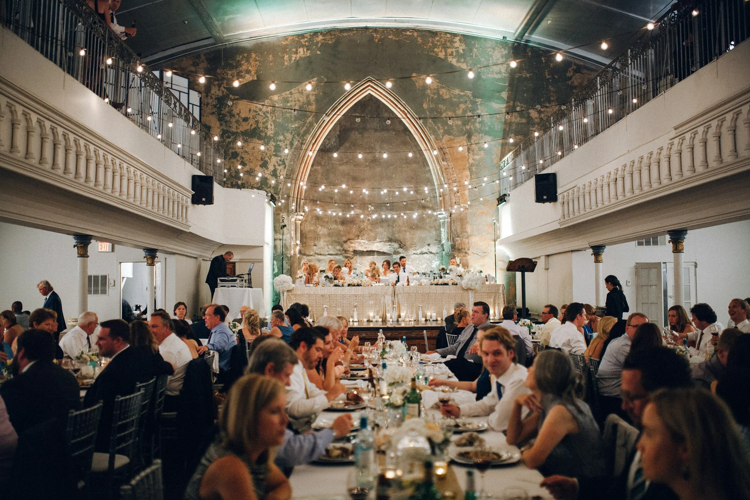 A large wedding reception in a spacious hall with rustic, worn green walls and decorative string lights hanging from the ceiling. Guests are seated at tables adorned with white floral centerpieces, enjoying their meal and conversation. The head table is elevated with a long table where the wedding party is sitting, facing the guests.