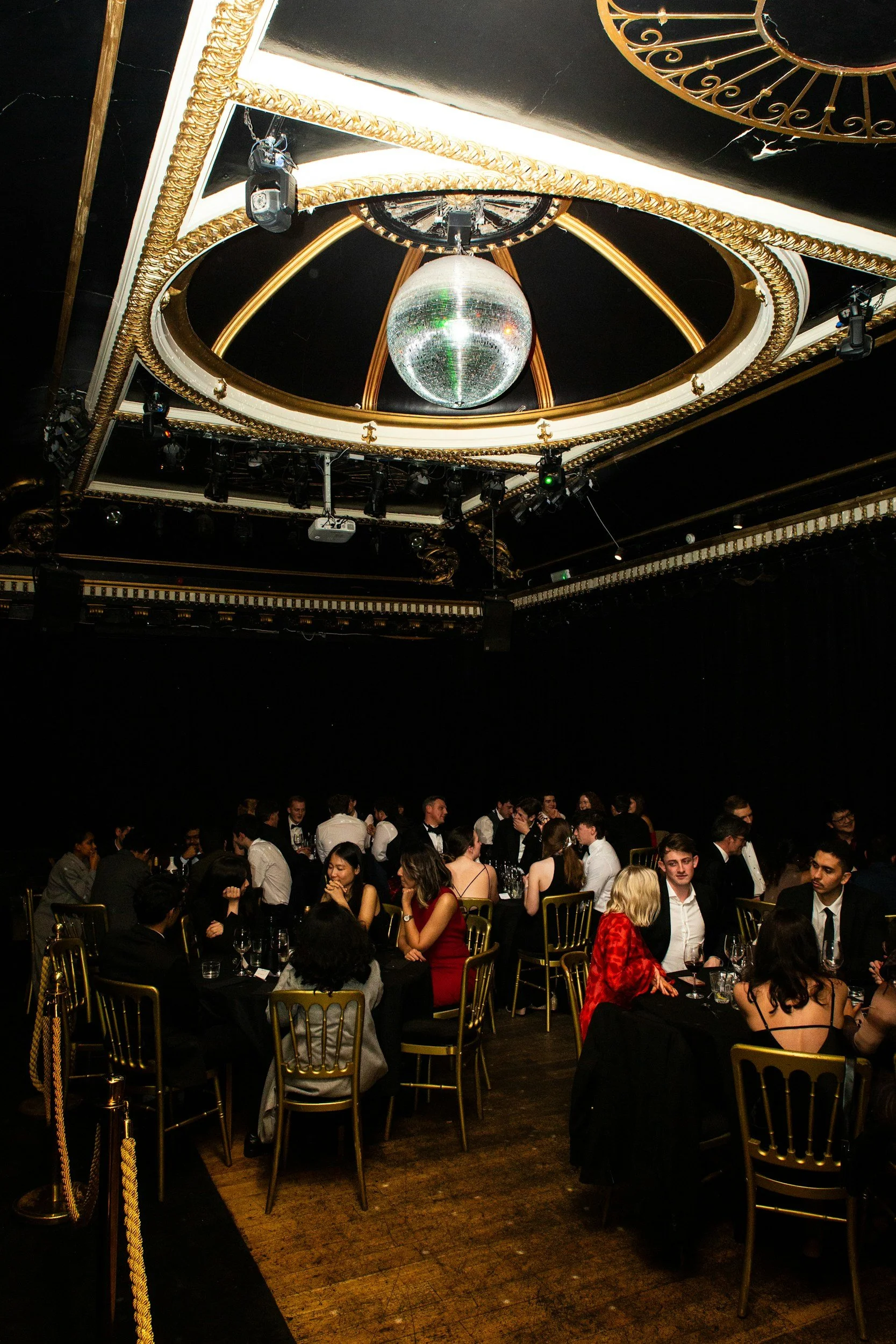 A large banquet hall with elegantly dressed guests seated at round tables with gold chairs. A disco ball hangs from the ornate ceiling, which features gold and white details, and stage lights are visible around the room.