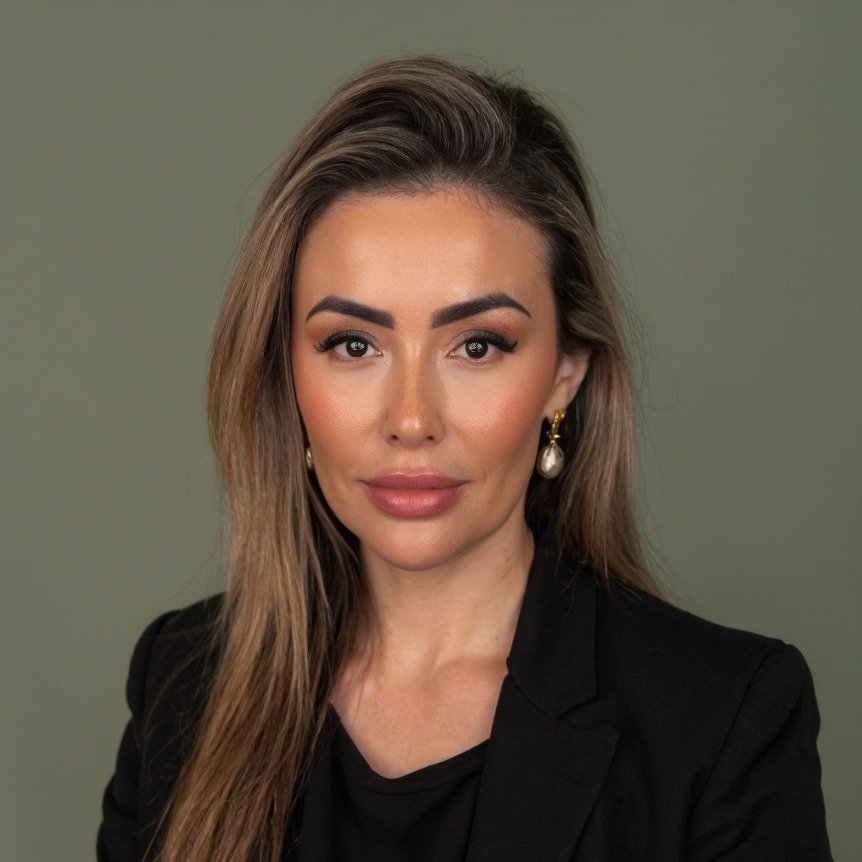 A woman with long, wavy light brown hair, wearing makeup, a black blazer, and pearl earrings, posing against a plain green background.