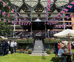 Outdoor stage decorated with British flags, surrounded by people, plants, and outdoor seating at a festive event.