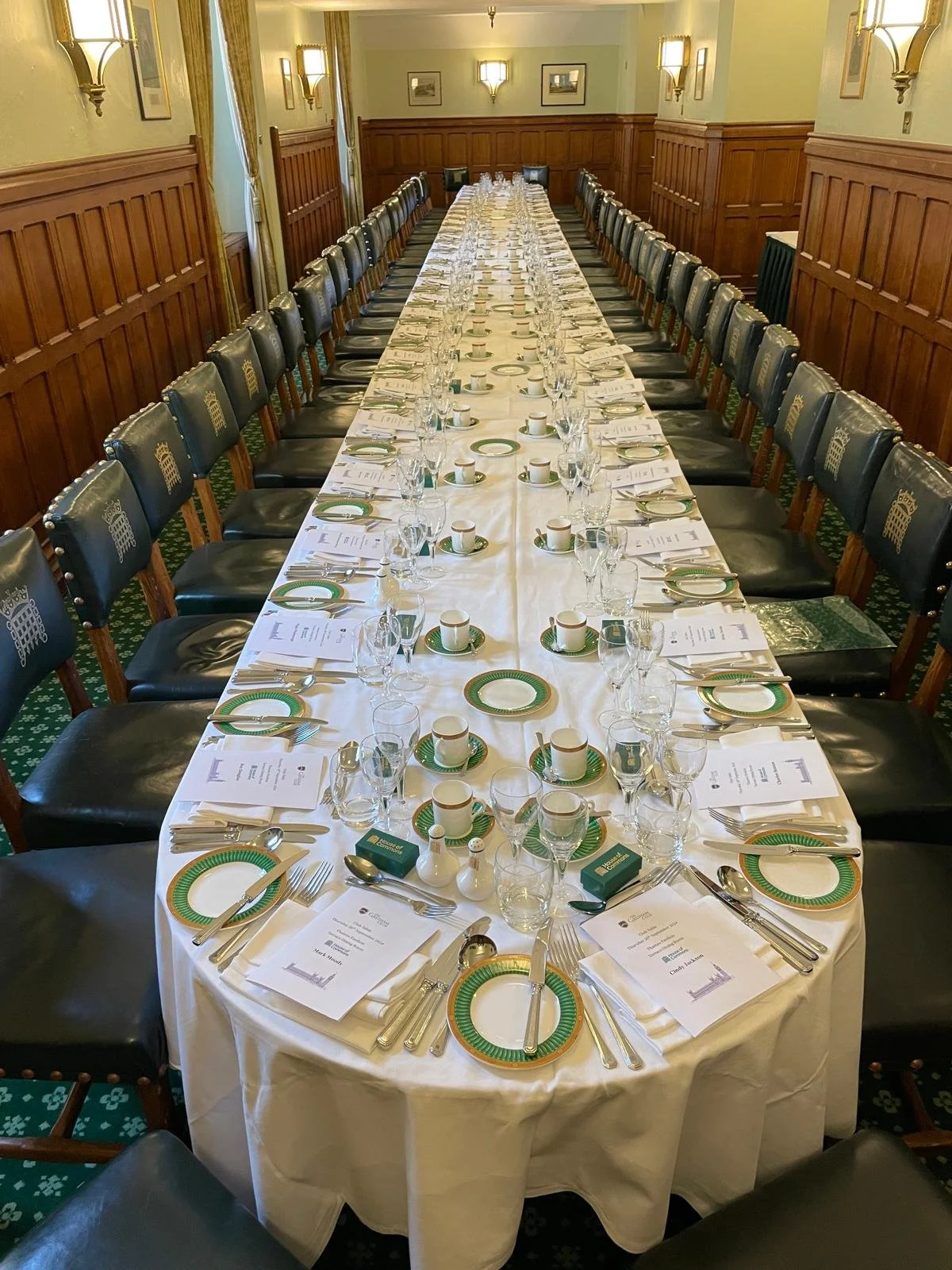 A long banquet table set for a formal meal in a wood-paneled room with green carpet and wall-mounted lights.