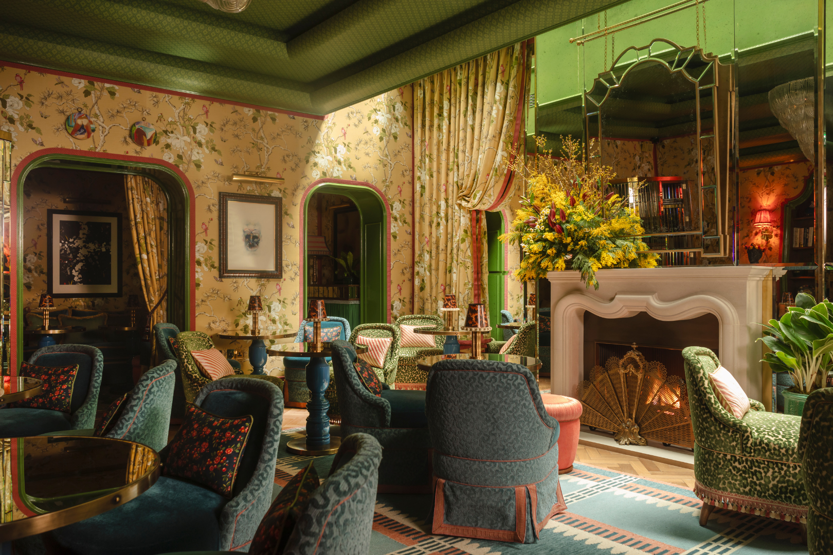 Interior of a vintage decorated lounge or sitting area with velvet armchairs, a white fireplace with a floral arrangement, patterned wallpaper, and mirror accents.