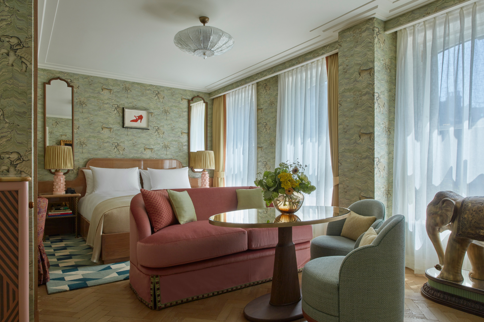 A hotel room featuring a bed with white pillows, pastel and patterned throw pillows, vintage-style lamps, a pink sofa with throw pillows, a round glass table with a flower vase, two green armchairs, and curtains covering large windows.