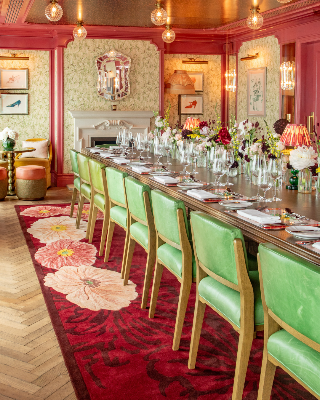 A fancy dining room set up for a meal with a long wooden table, green chairs, and floral centerpieces. The room has patterned wallpaper, framed artwork, pink molding, and a colorful floral rug. There are glasses, plates, and silverware arranged on the table.