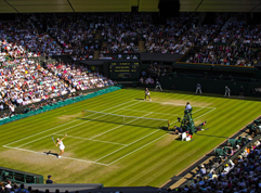 A professional tennis match on a grass court with players, a umpire, and a line judge, surrounded by spectators in a stadium.
